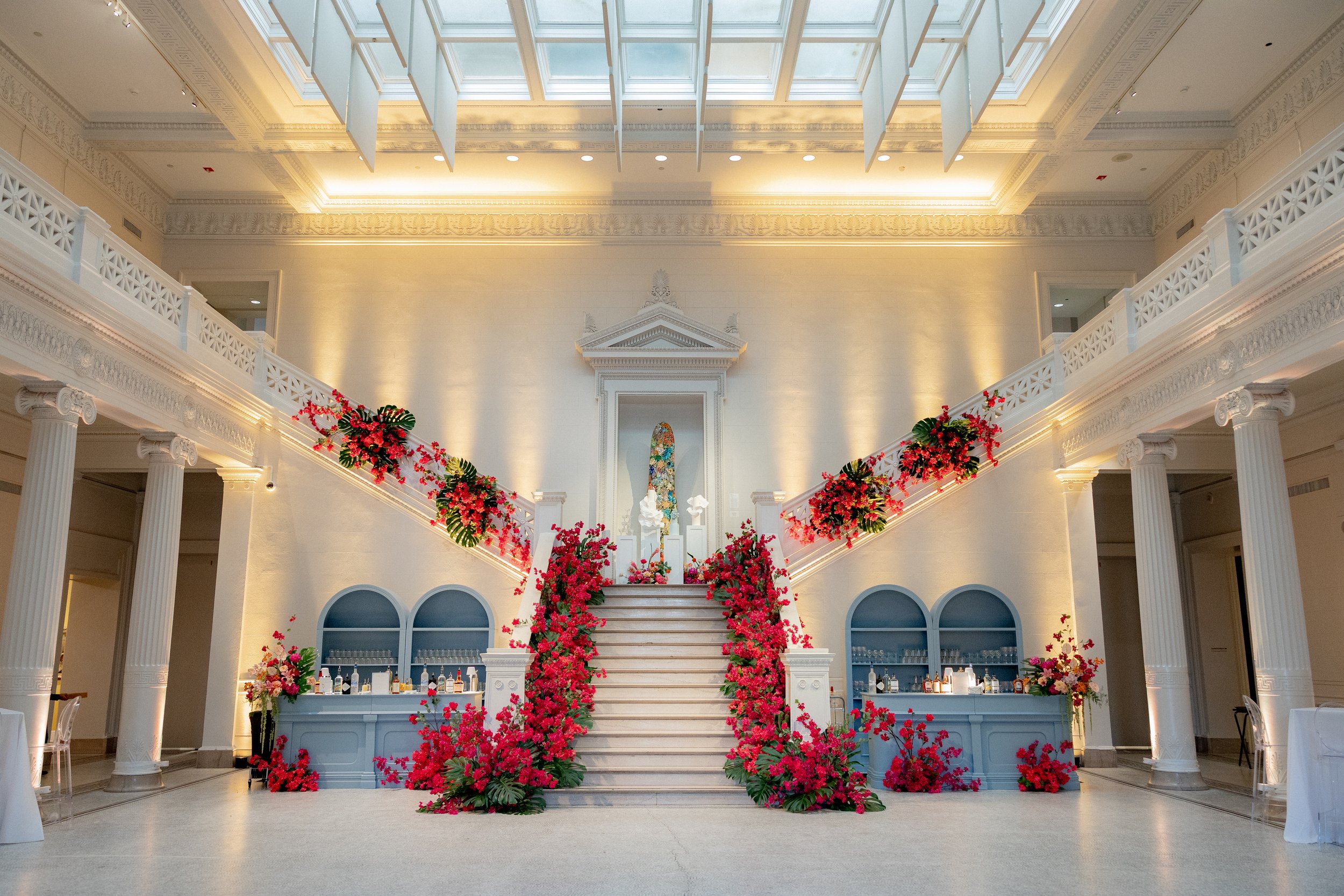 Interior view of a grand hall with white walls and columns, decorative molding, large skylight, and floral decorations on staircase and surrounding areas.