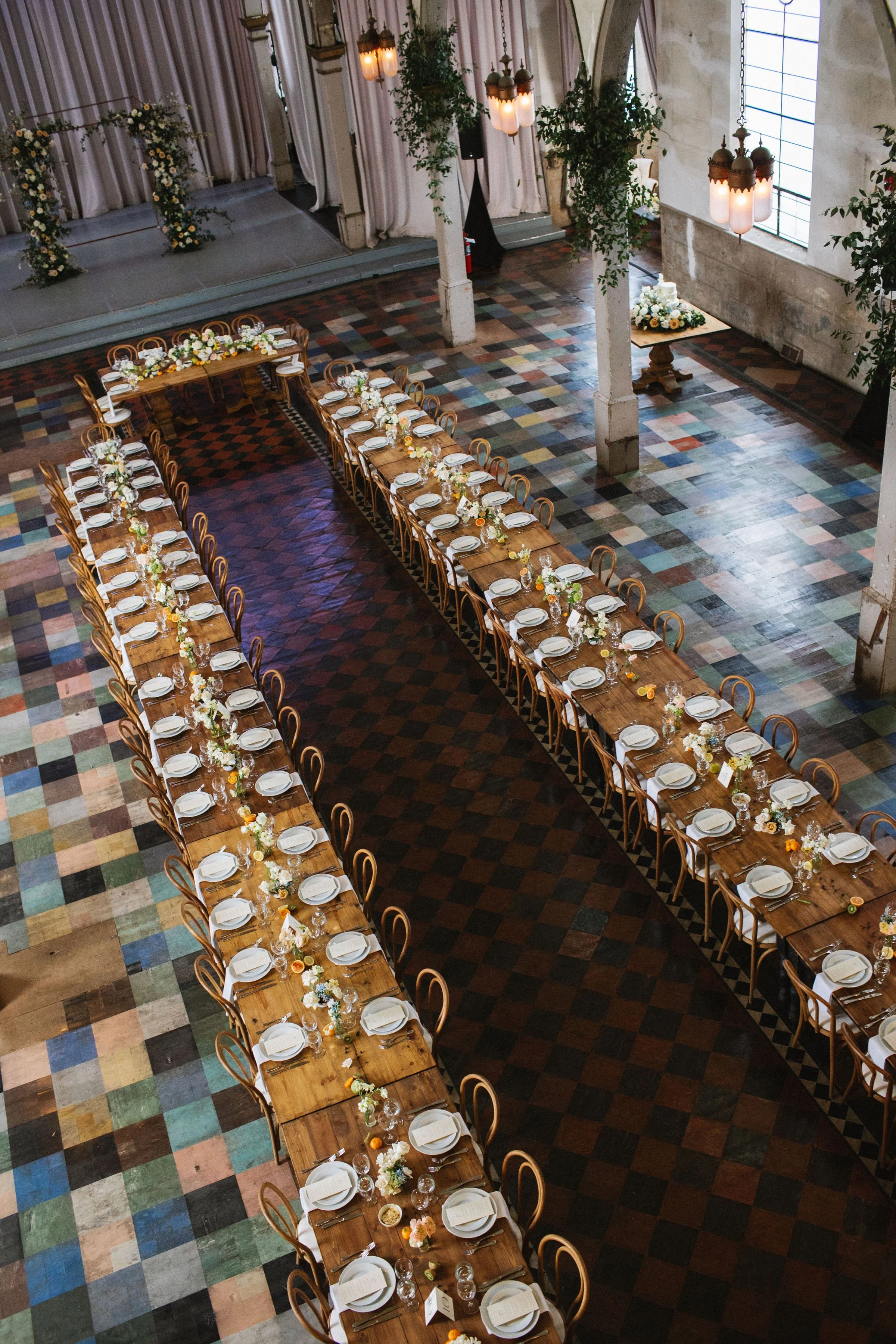 Long wooden banquet tables set with white plates, glasses, utensils, and small flower arrangements, in an industrial-style venue with high ceilings, large windows, and hanging lanterns, likely prepared for a wedding or formal event.