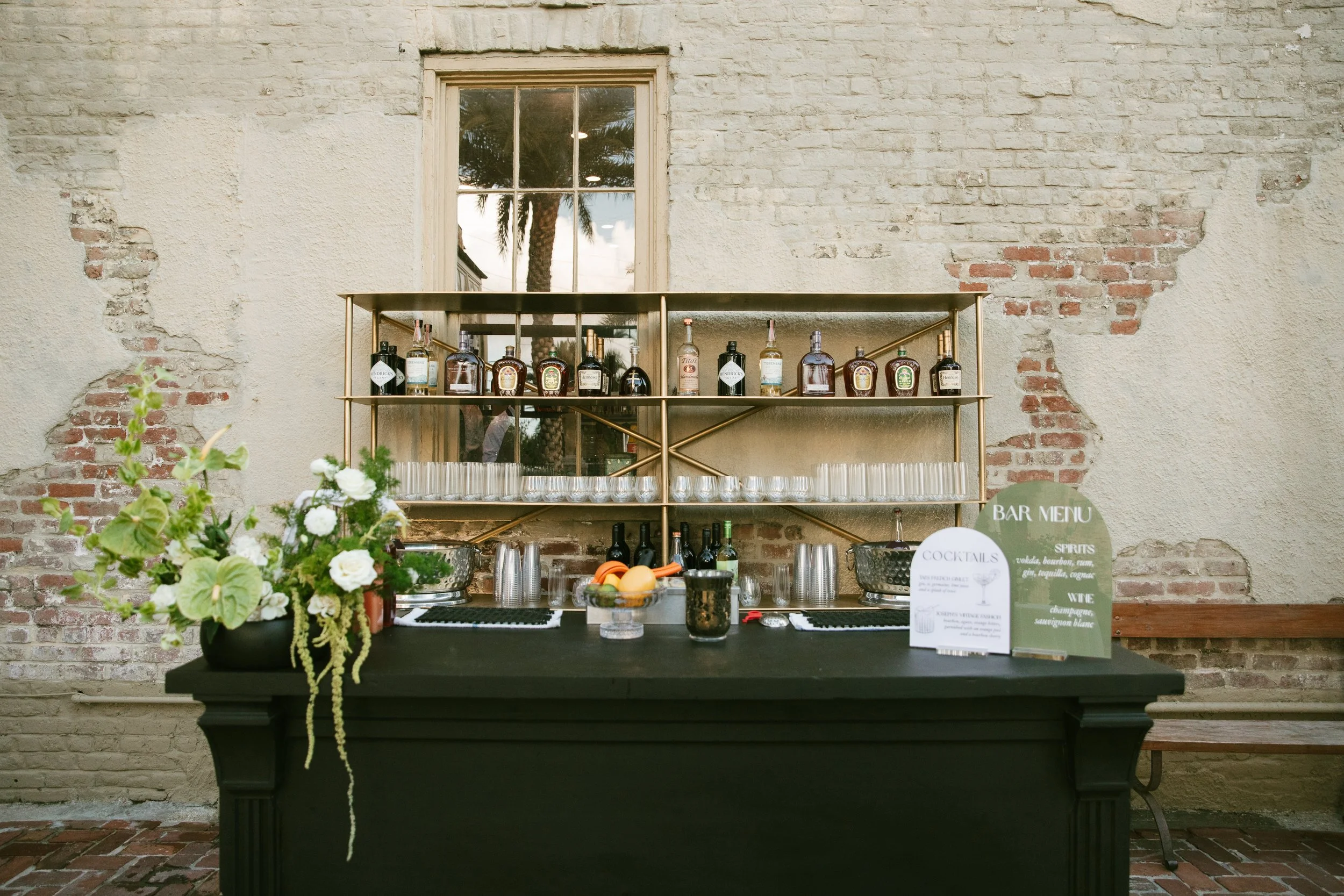 Bar area with bottles of liquor on a gold metal shelf, floral arrangement on a black table, and two menu signs, against a brick wall with a window reflecting palm trees.