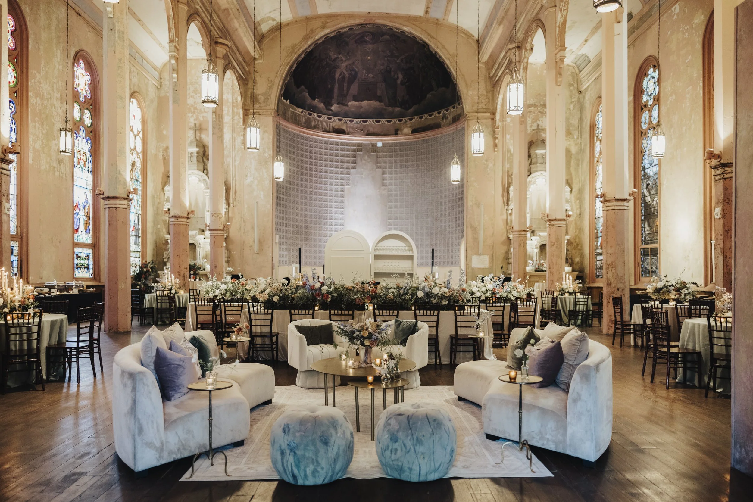 An elegantly decorated interior of a church or cathedral with stained glass windows, ornate columns, and chandeliers, set up for a wedding or reception with round tables, floral centerpieces, and cushioned seating.