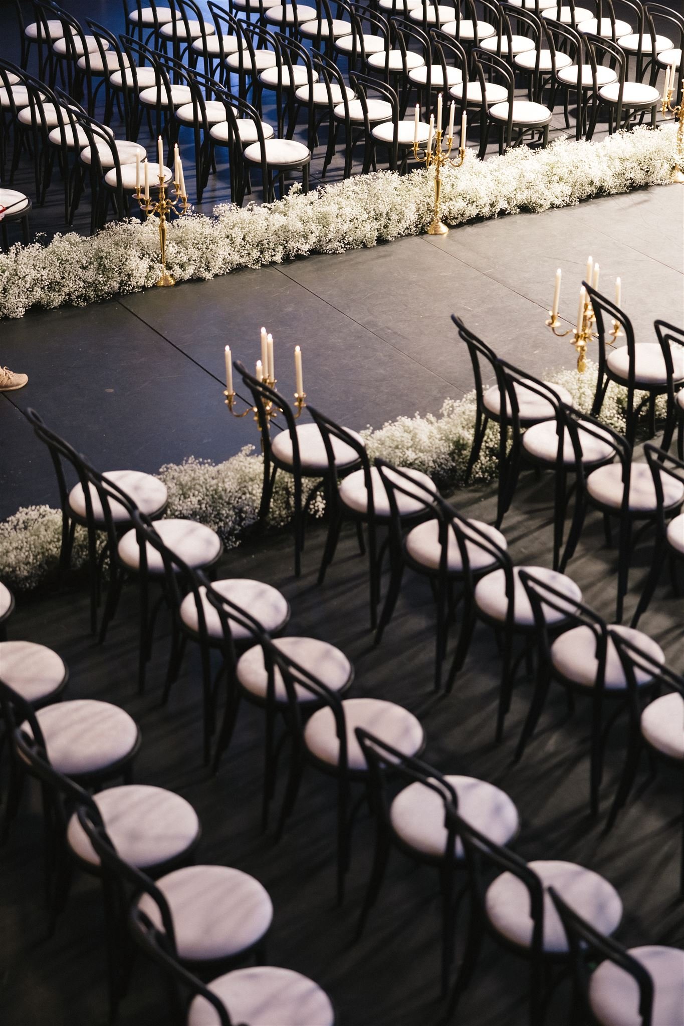 Empty black chairs with white cushions arranged in rows for a wedding ceremony, lined with white baby's breath flowers, gold candle holders, and candles on a dark floor.
