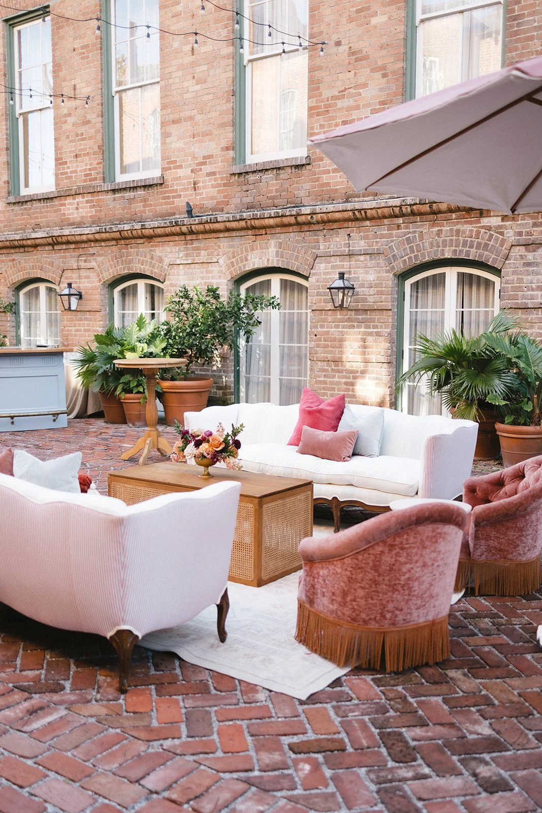 A cozy outdoor patio with vintage furniture, including a white sofa, pink velvet chairs, and a wooden coffee table with a floral arrangement. Potted plants and brick walls with windows and string lights surround the space, creating a charming, relaxe