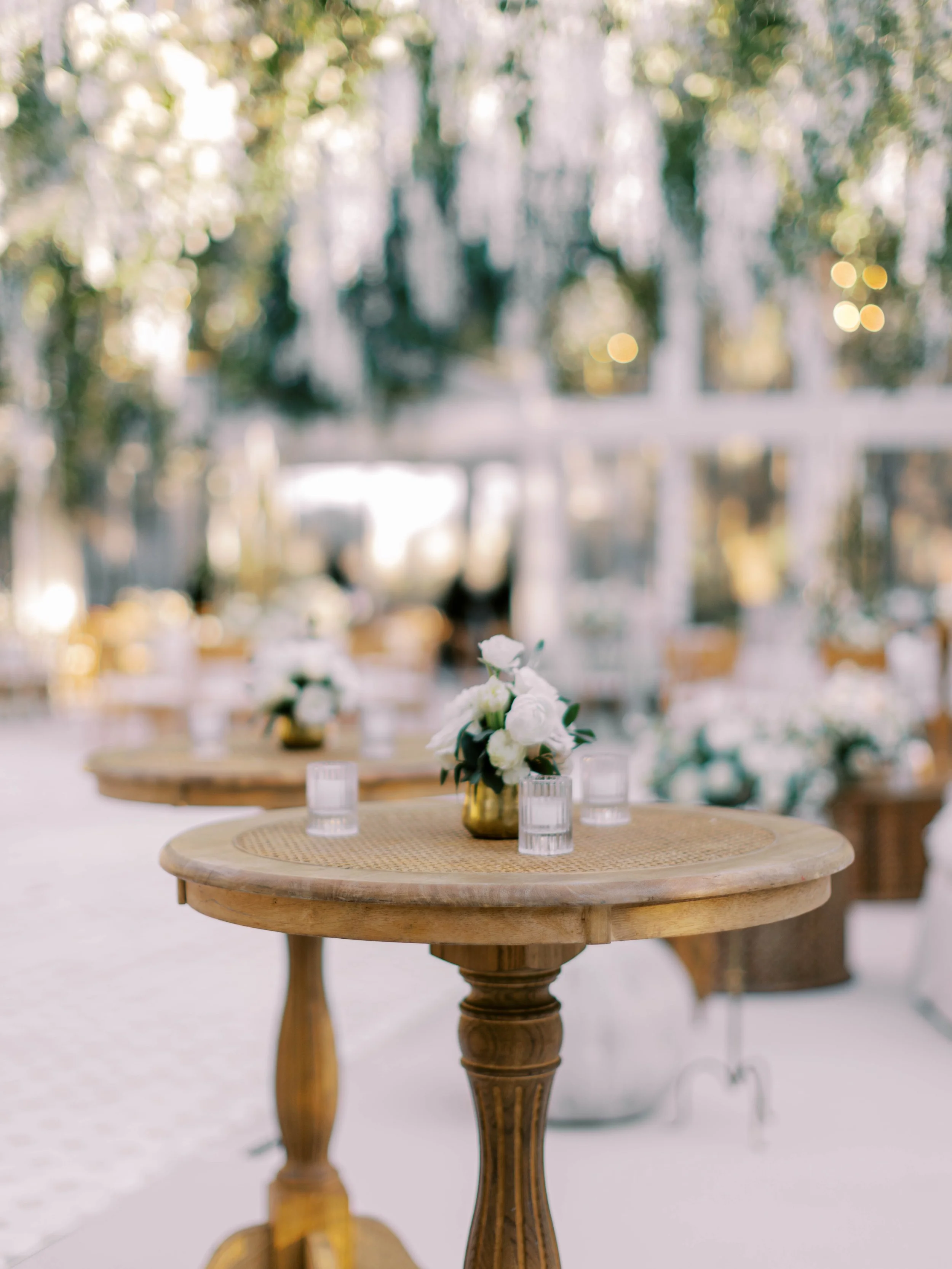 A round wooden table with a flower arrangement in a gold vase and several small candles, with blurred background of trees and sunlight.