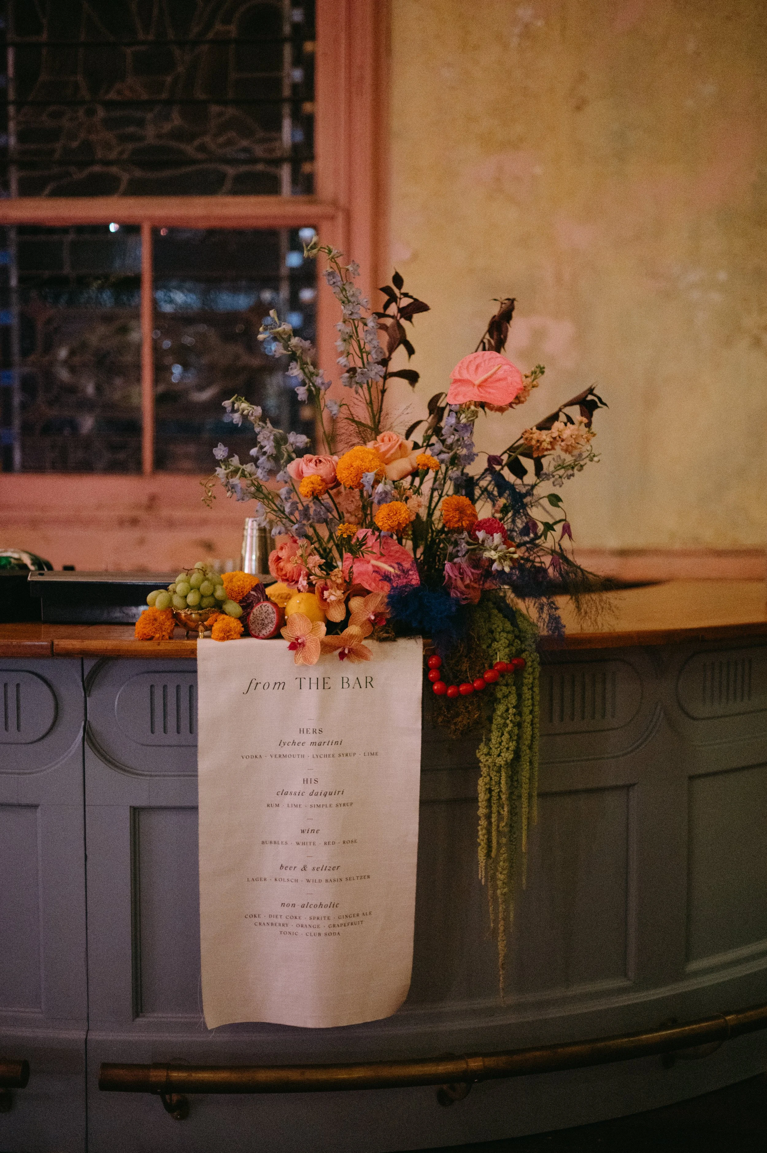 A floral arrangement on a bar counter with a printed list of drinks attached below. The arrangement includes pink, purple, orange, and yellow flowers with green foliage. The background features a window with a stone wall outside and a textured yellow
