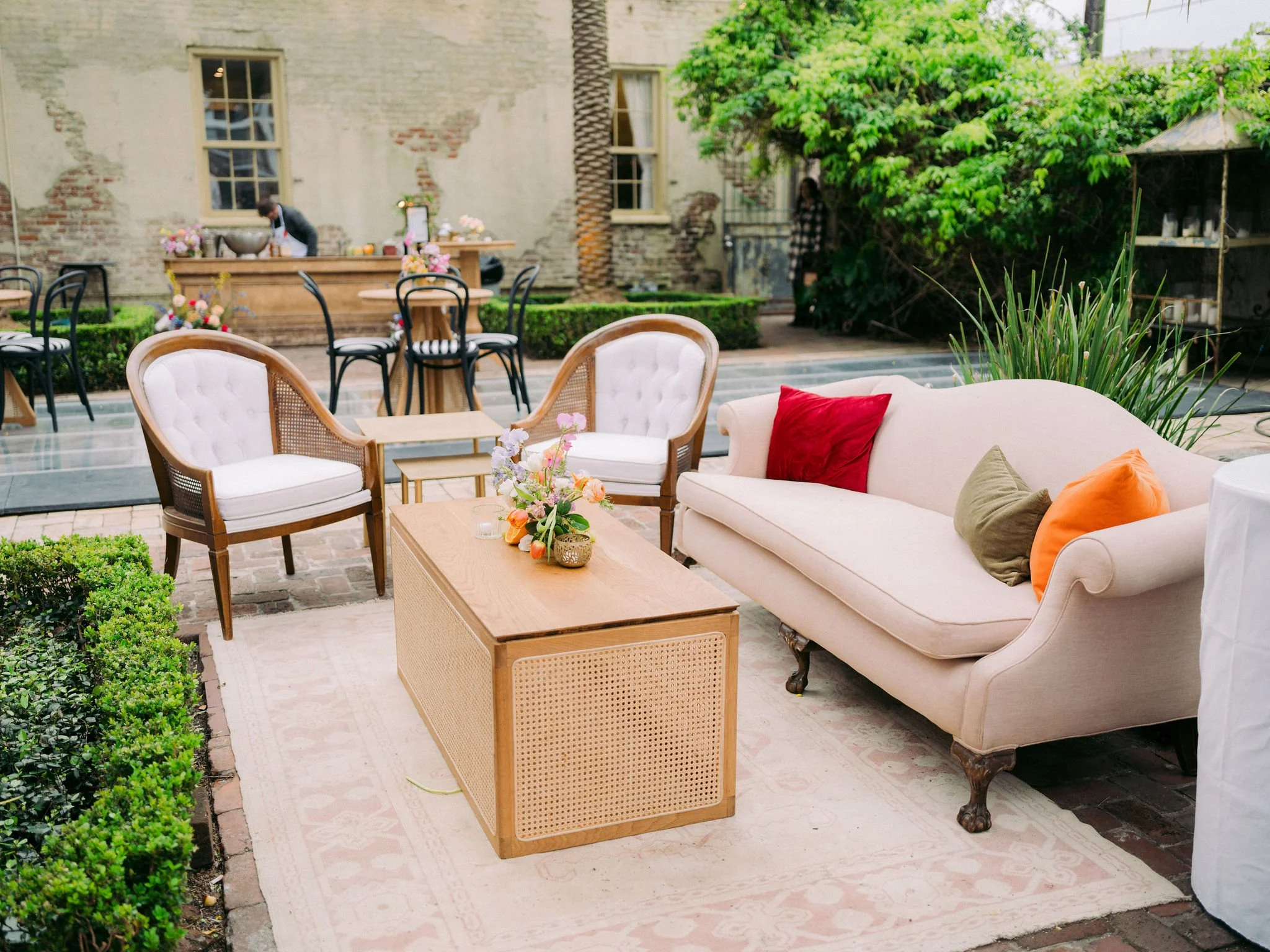An outdoor patio area with vintage-style furniture, including a beige sofa with colorful cushions, armchairs, and a wooden coffee table with a floral centerpiece. In the background, there are black chairs around a wooden table, greenery, trees, and a