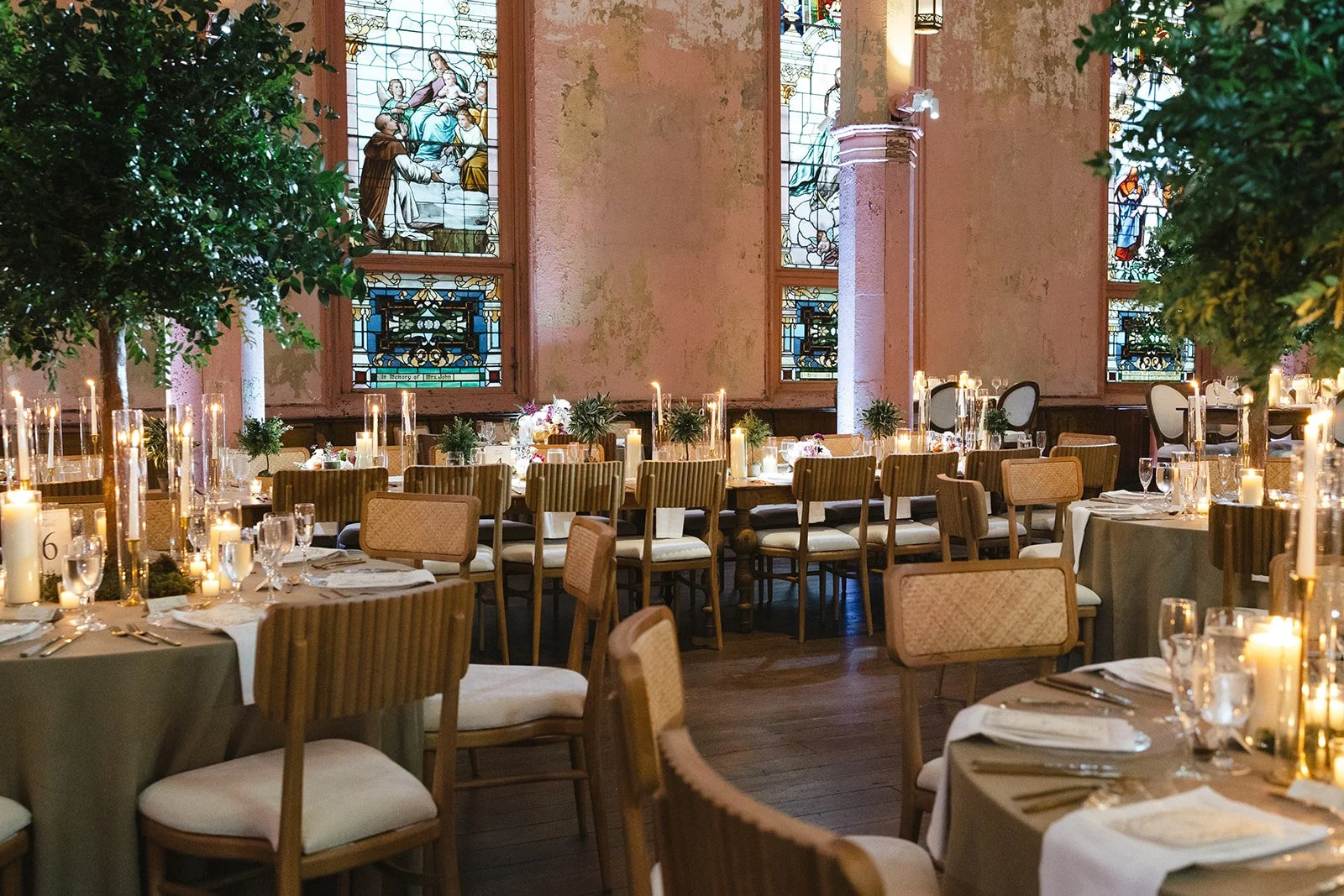 Elegant banquet hall with round tables covered in beige tablecloths, surrounded by wooden chairs with woven backs, set with glassware, silverware, and white napkins, decorated with candles and greenery, large stained glass windows depicting religious