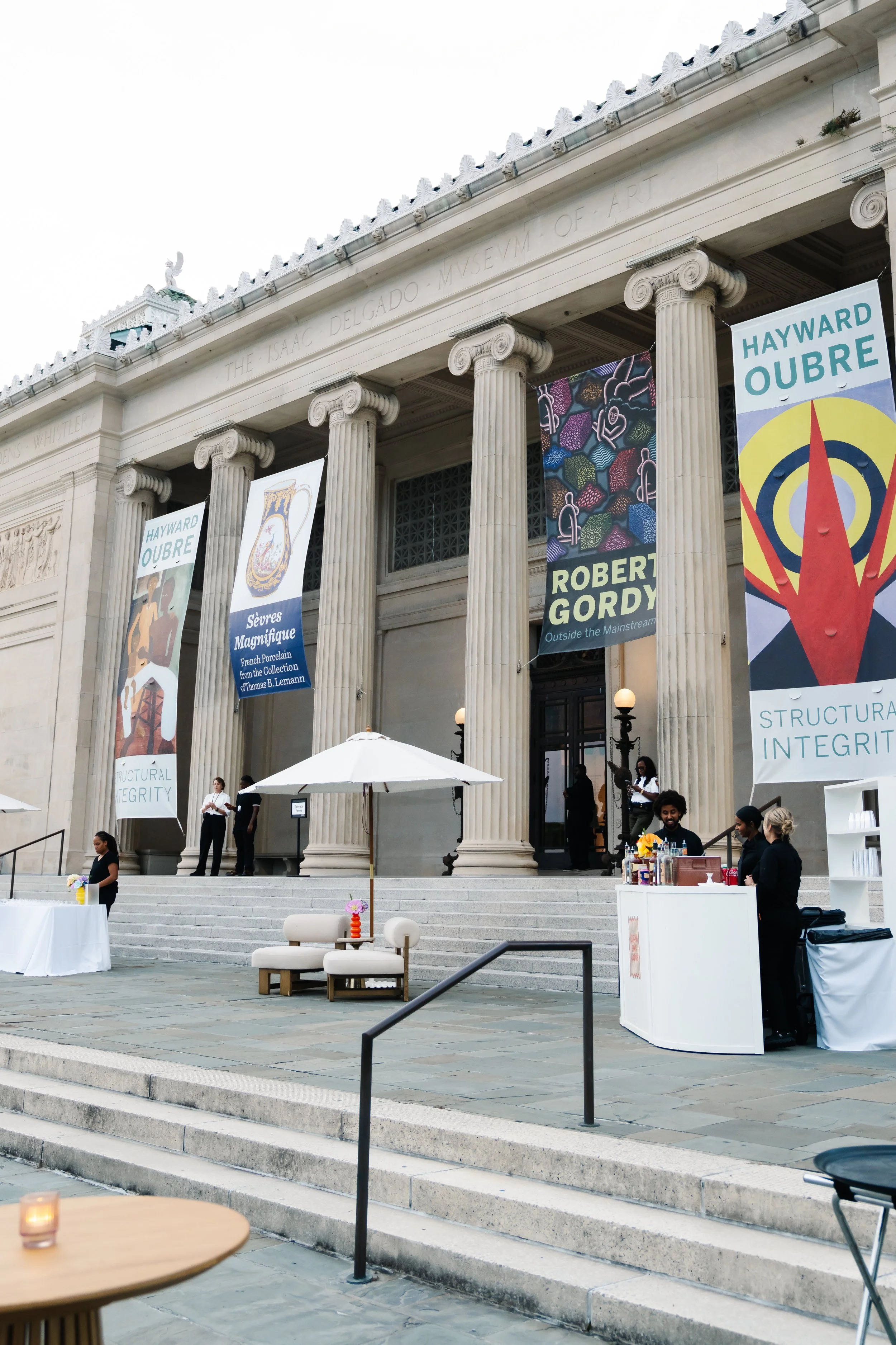 The entrance of the Isaac Delgado Museum of Art with large columns and banners advertising exhibitions by Hayward Oubre, Robert Gordy, and Sévres Magnifique. There are steps leading up to the entrance, and people are gathered on the steps and around 