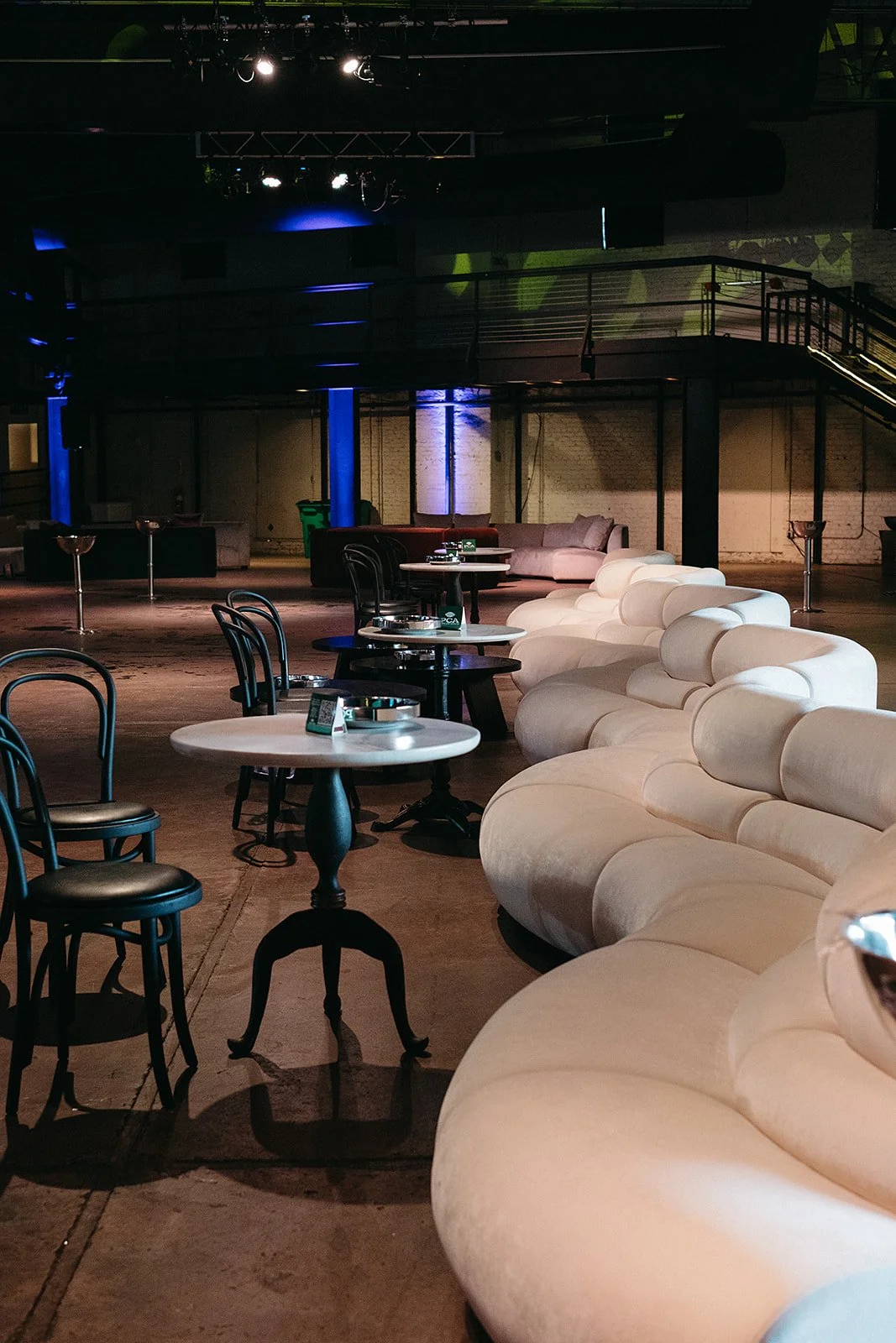 Empty lounge with white curved sofas, black chairs, and round tables in a dimly lit space, possibly a nightclub or bar.
