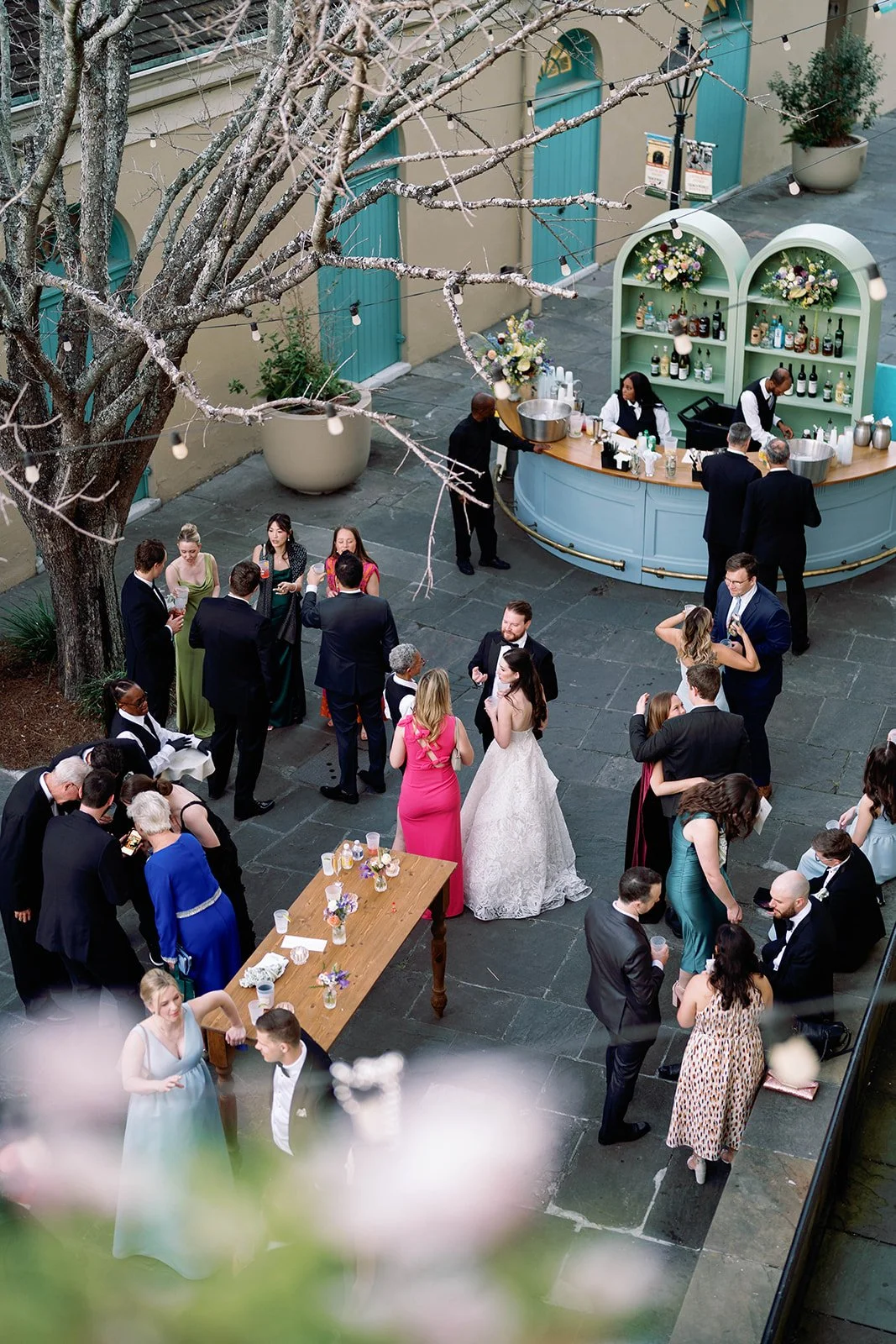 A wedding reception in an outdoor courtyard with guests mingling around a bar and a decorated table, a large tree, string lights, and potted plants.
