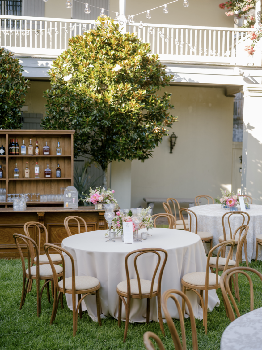 Outdoor event setup with round tables, beige chairs, floral centerpieces, a bar with liquor bottles, and string lights on a lawn.
