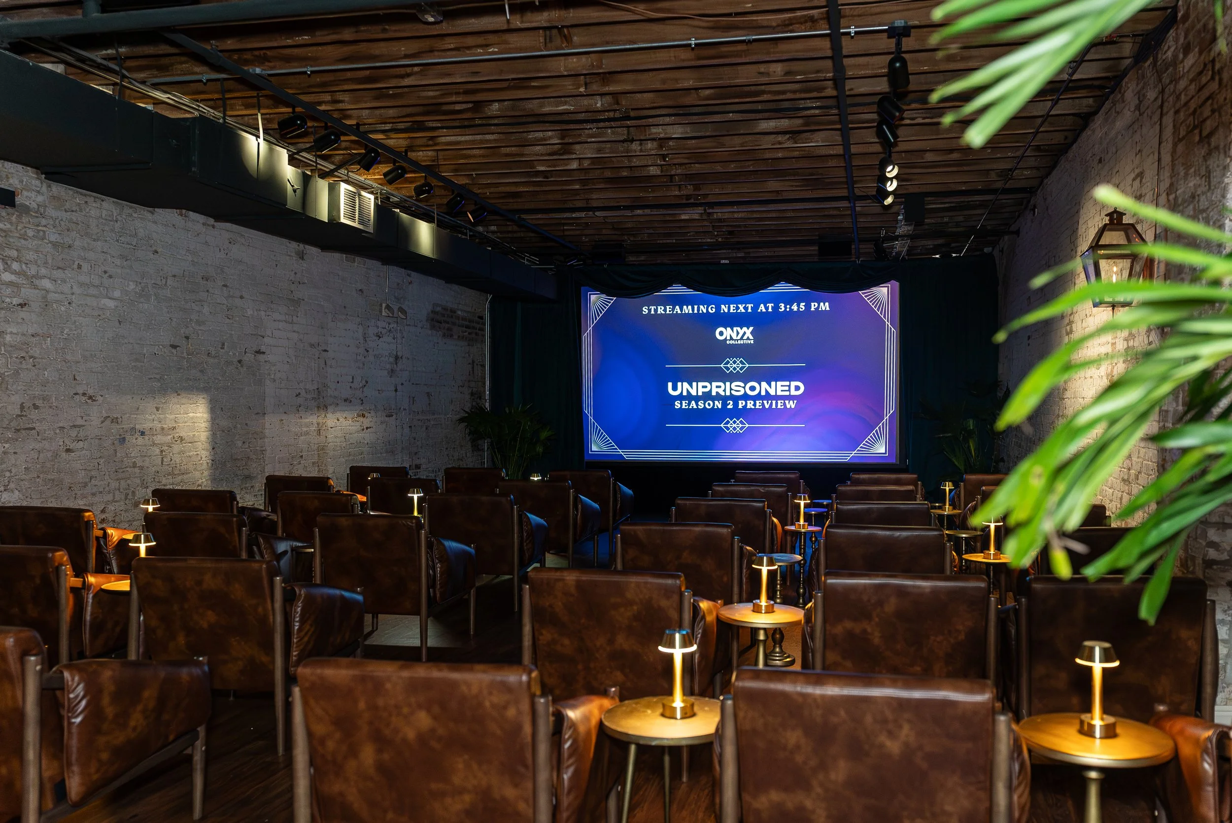 An indoor event space with rows of brown leather chairs, small tables with lamps, a large digital screen displaying information about a streaming event, brick walls, a wooden ceiling, and some green plants in the foreground.