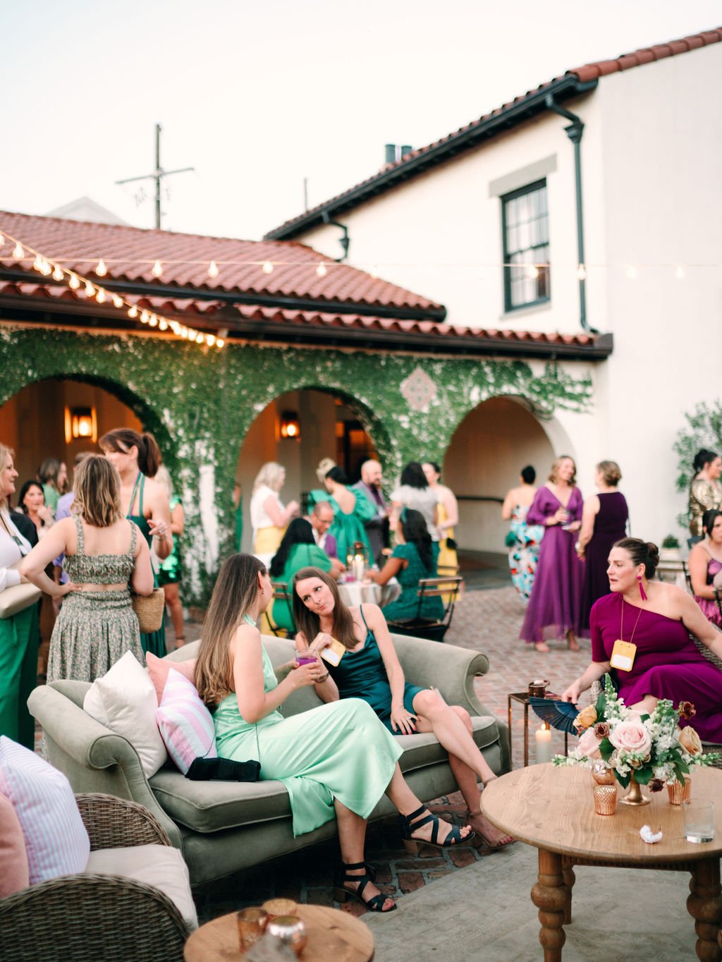 People socializing at an outdoor gathering with couches, a table with flowers, string lights, and a building covered in green foliage in the background.