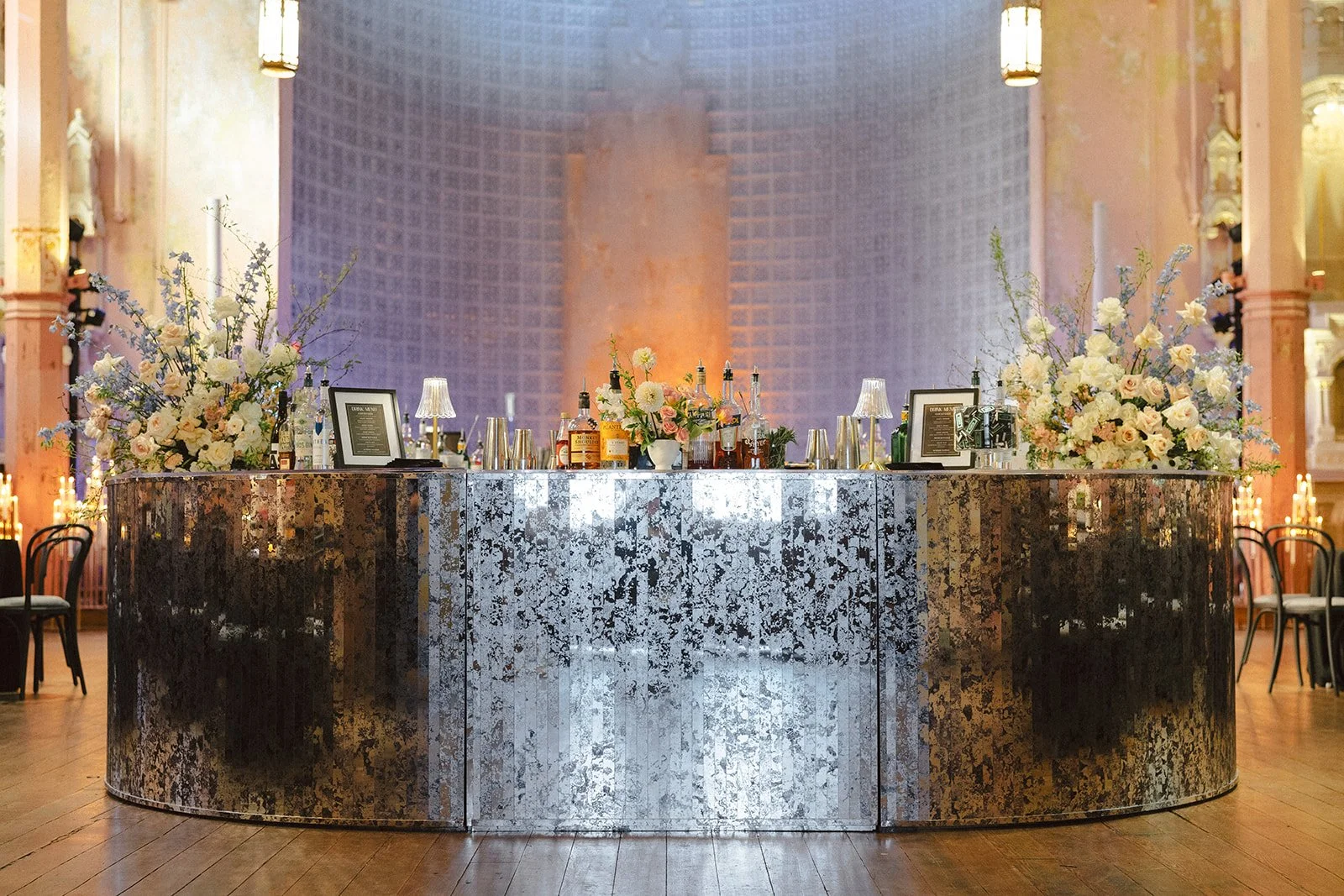 Elegant bar setup with floral arrangements, framed menus, liquor bottles, and small lamps, in a luxurious event space with high ceilings and ornate decor.