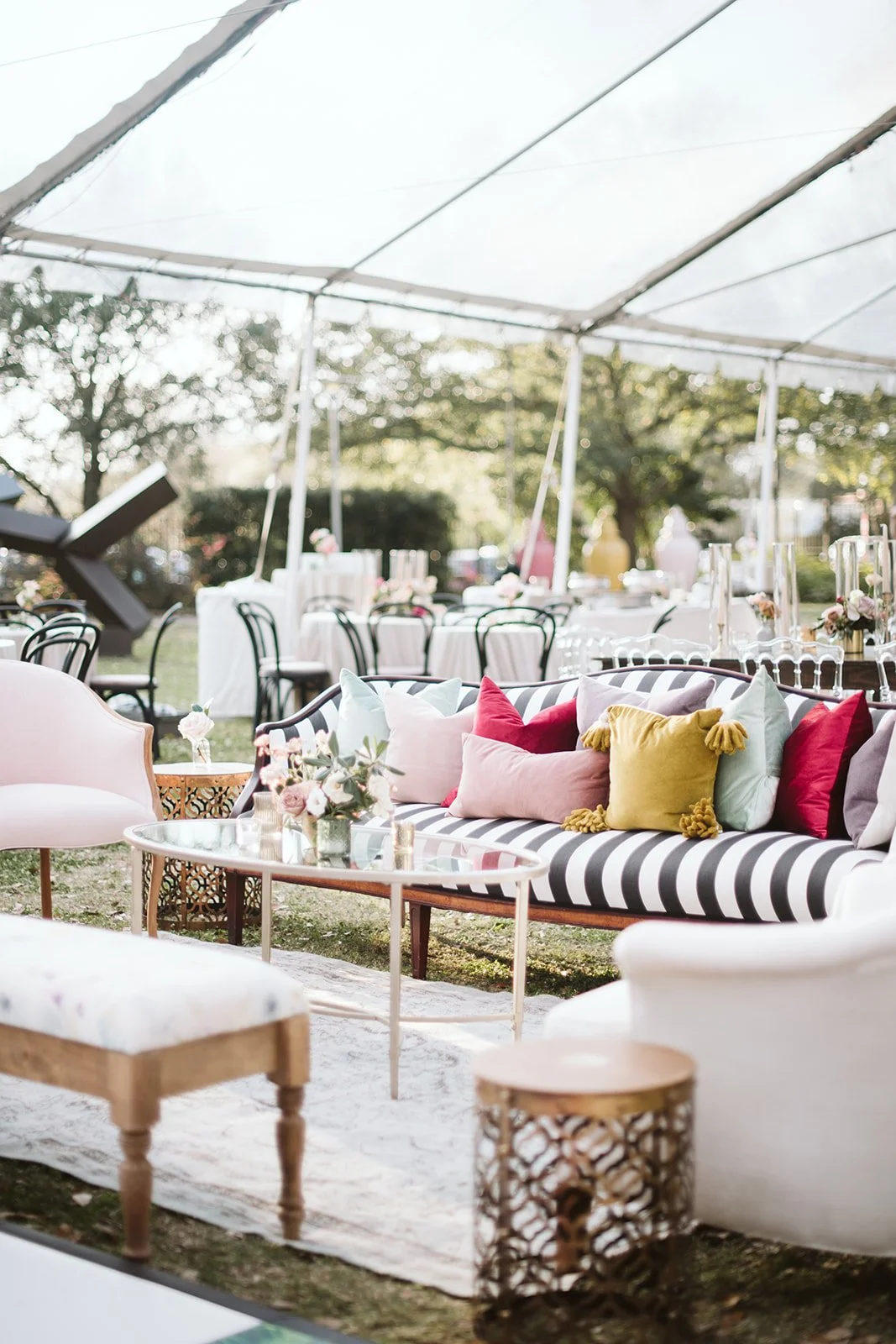 Decorated outdoor event space with a striped sofa, colorful pillows, white tables, chairs, flowers, and a canopy tent in the background.