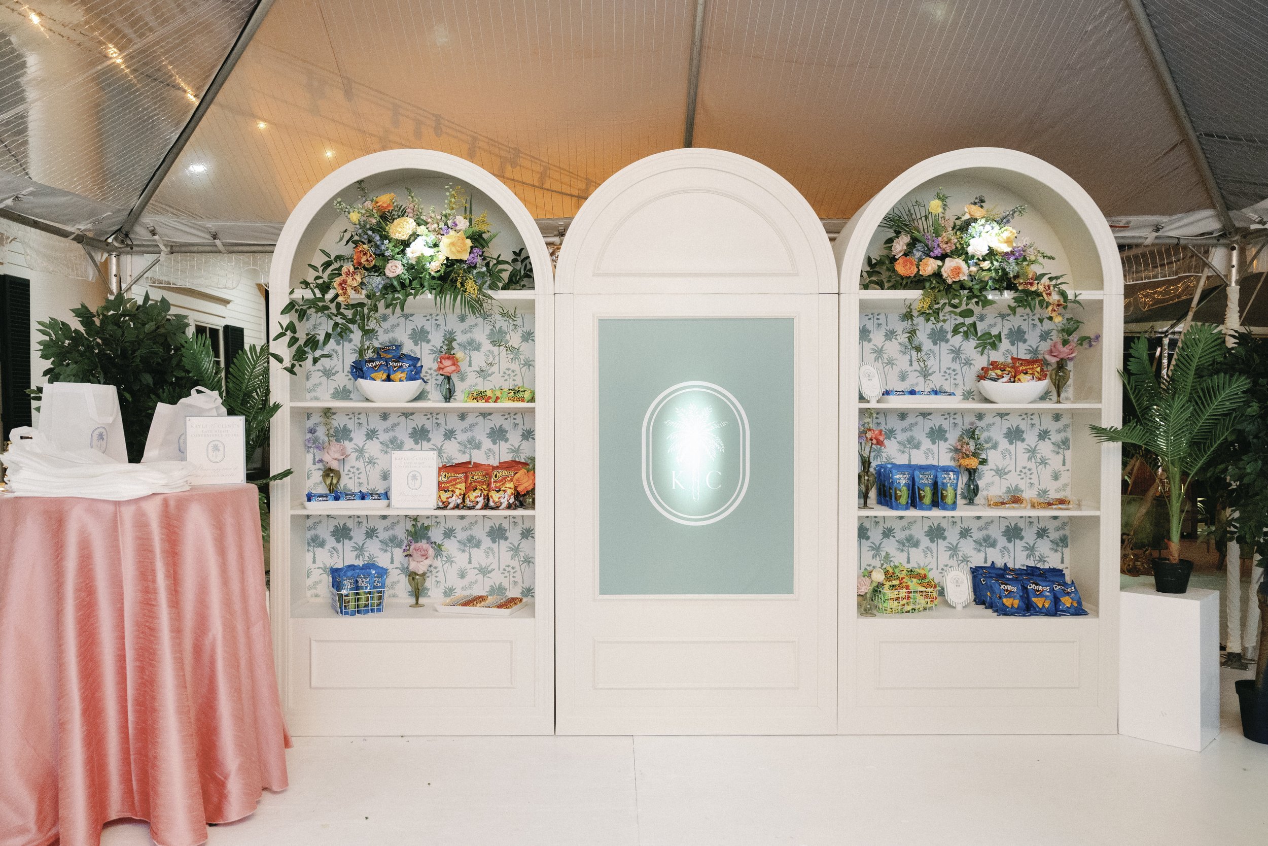 White decorative shelving unit with floral arrangements and snack bags, flanked by potted plants, in a tented indoor space.