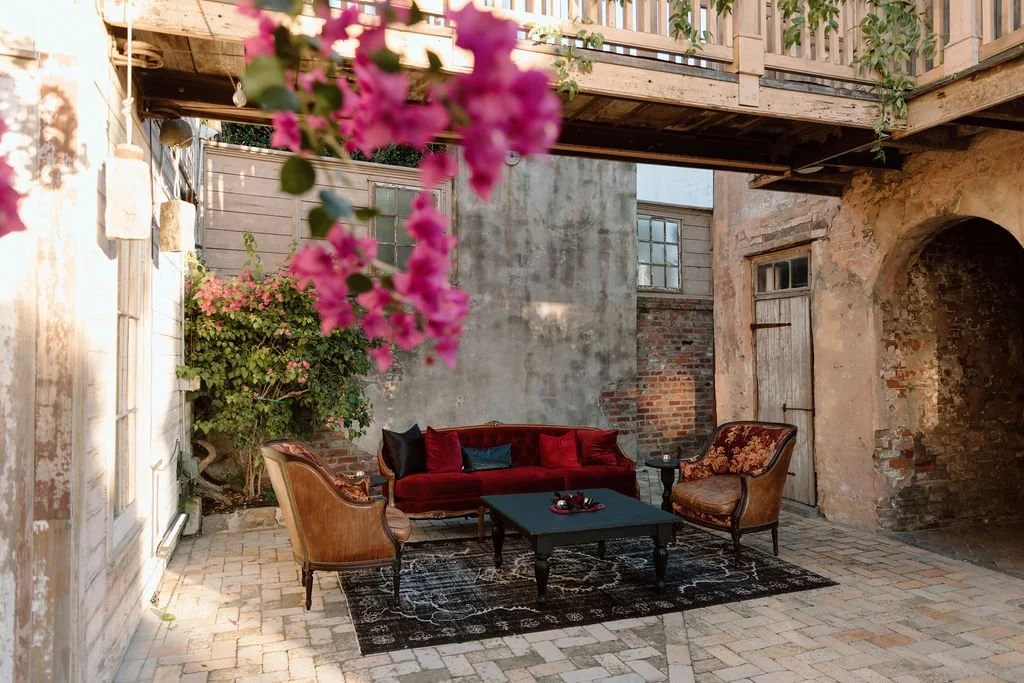 Outdoor patio or courtyard with vintage furniture, including a red velvet sofa, two armchairs, a black coffee table, and decorative pillows, surrounded by brick and stone walls, plants, and pink flowers hanging in the foreground.
