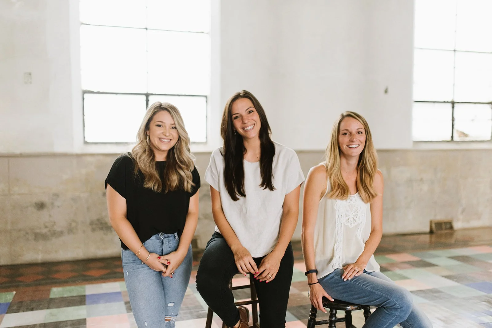 Three women smiling and sitting on stools in front of large windows in a bright, industrial-style room with tiled floor.
