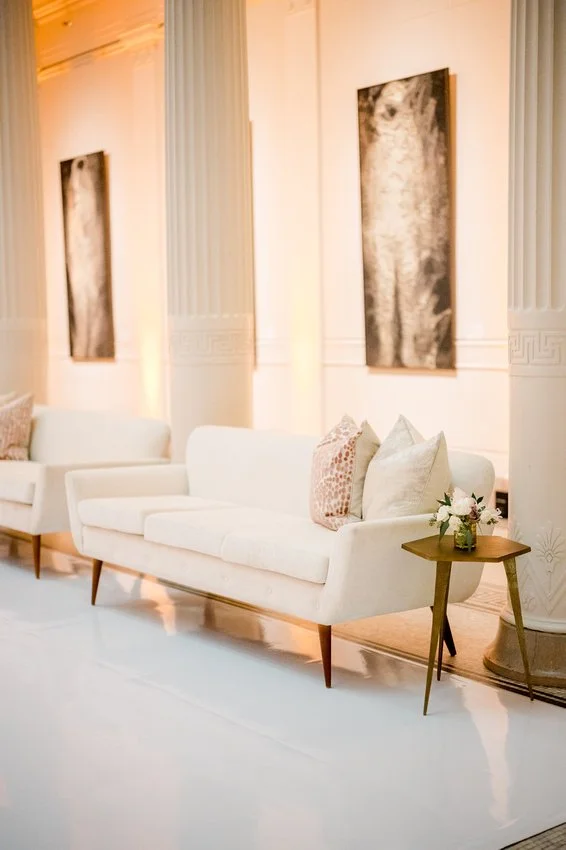 A bright, elegant living room with white sofas, decorative pillows, a small side table with flowers, and abstract artwork on cream-colored walls.