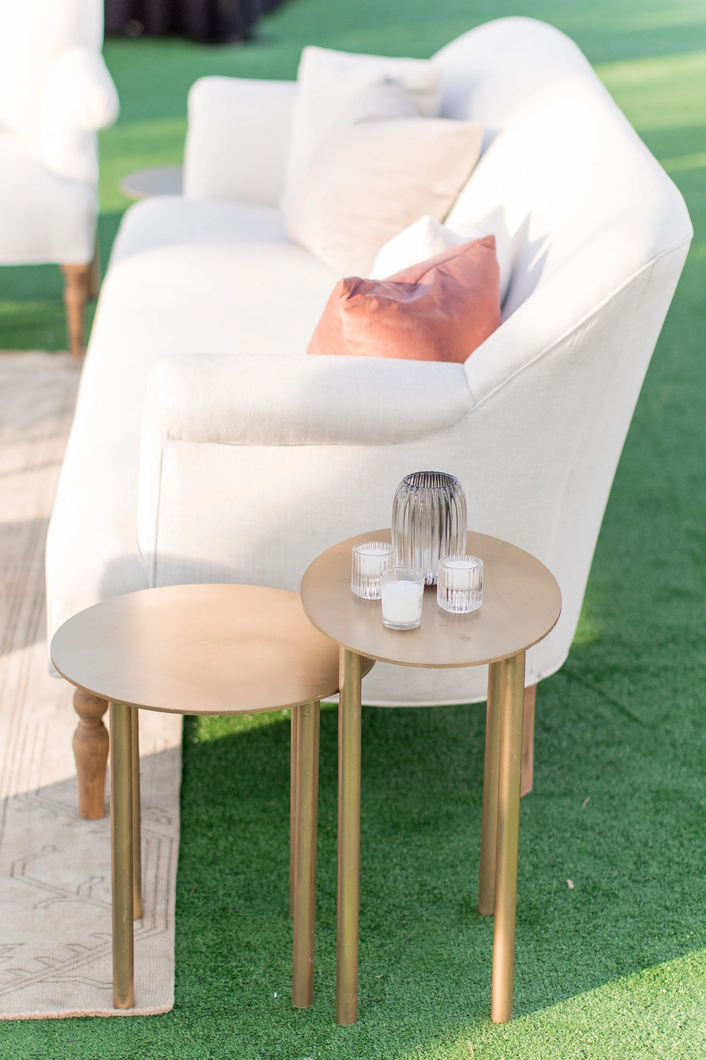 Outdoor patio setup with a white armchair decorated with beige, white, and rust-colored pillows. Two small round gold-colored side tables hold glassware, set on green artificial grass.