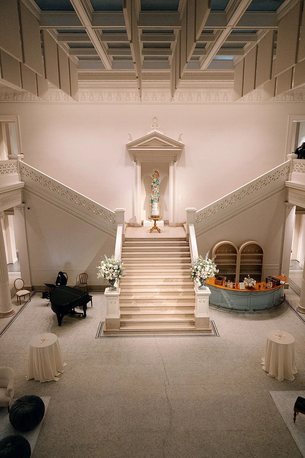 Elegant interior of a grand hall with a staircase, floral arrangements, a piano, and a bar area. Decorative architecture and a religious statue are visible.