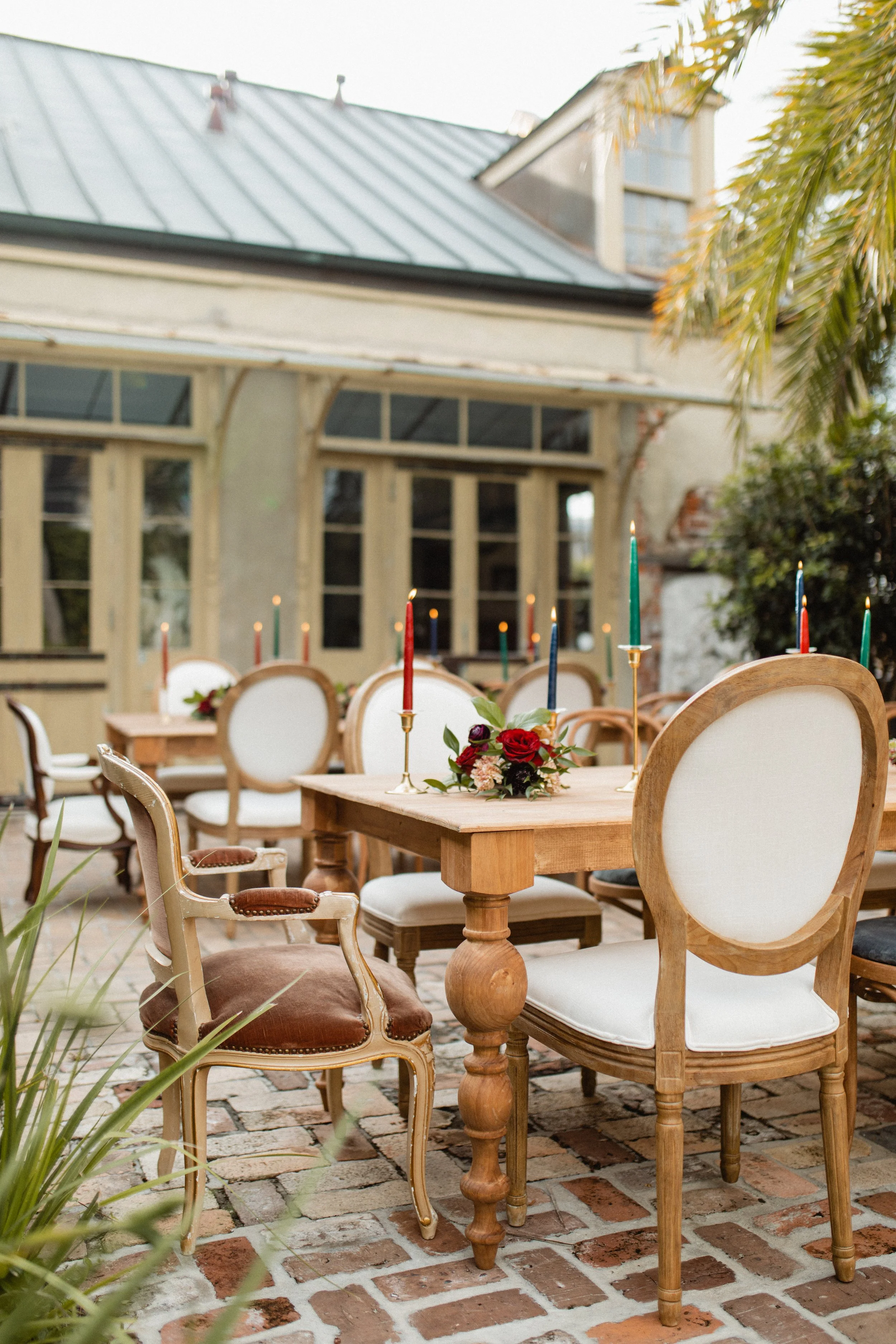 Outdoor patio with vintage and modern chairs around a wooden table decorated with candles and flowers, in front of a house with large windows and a metal roof, brick paving on the ground, and trees and plants nearby.