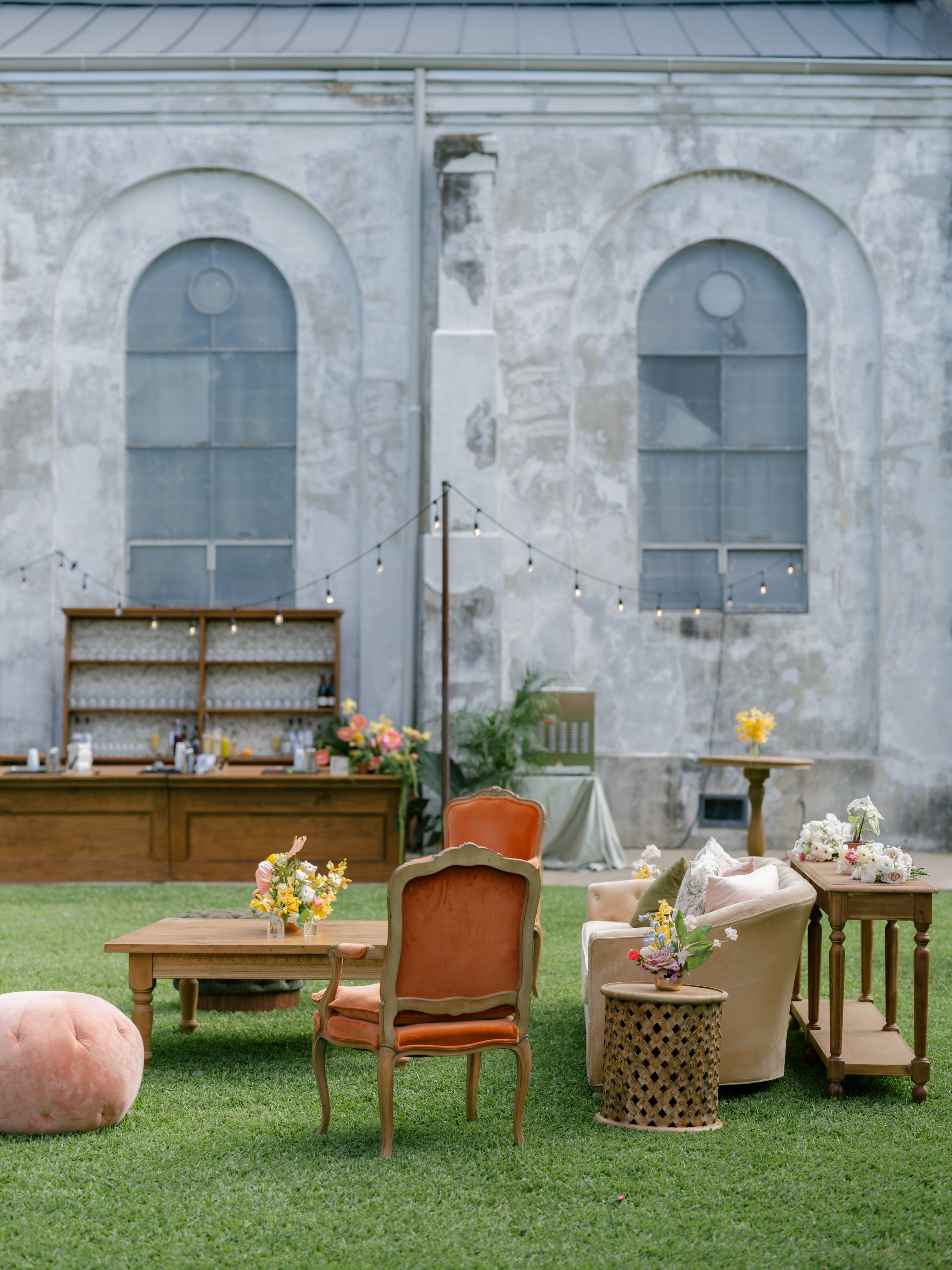 Outdoor wedding reception setup with vintage chairs, floral arrangements, string lights, and a rustic bar against a weathered building wall.