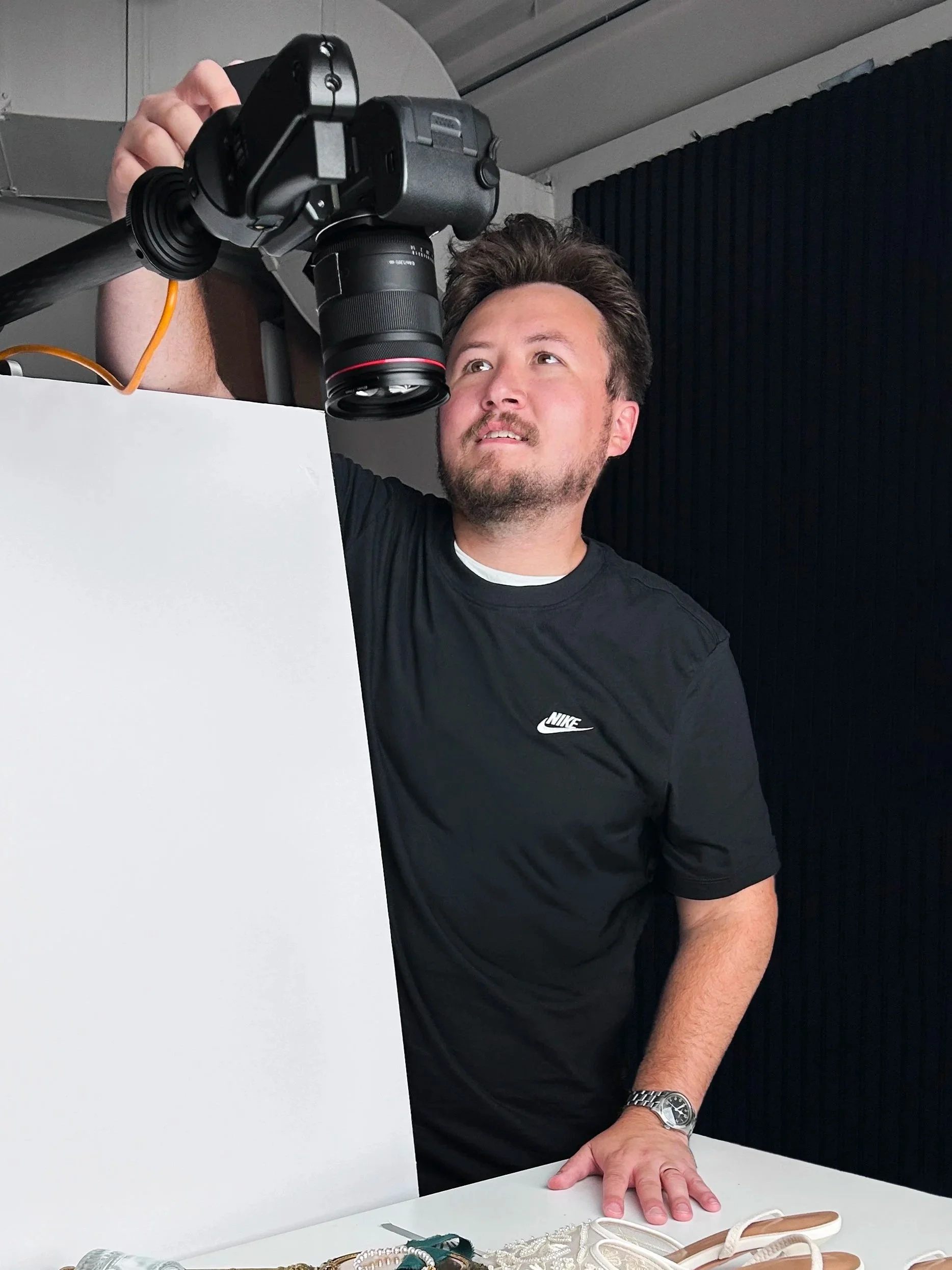 A man with brown hair, a beard, wearing a black Nike shirt and a silver wristwatch, is taking a photograph with a camera from a high angle. He is leaning on a white table with various jewelry items and a pair of beige sandals placed on it. The background features a black curtain and a gray ceiling.