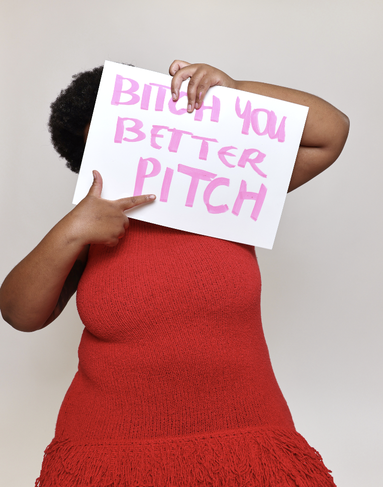 Woman in red dress holding 'B**** you better pitch' on white card