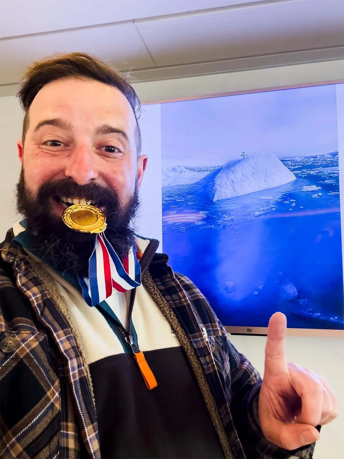 Tony J Hughes biting his gold medal in front of his prize winning photo of a penguin on an iceberg