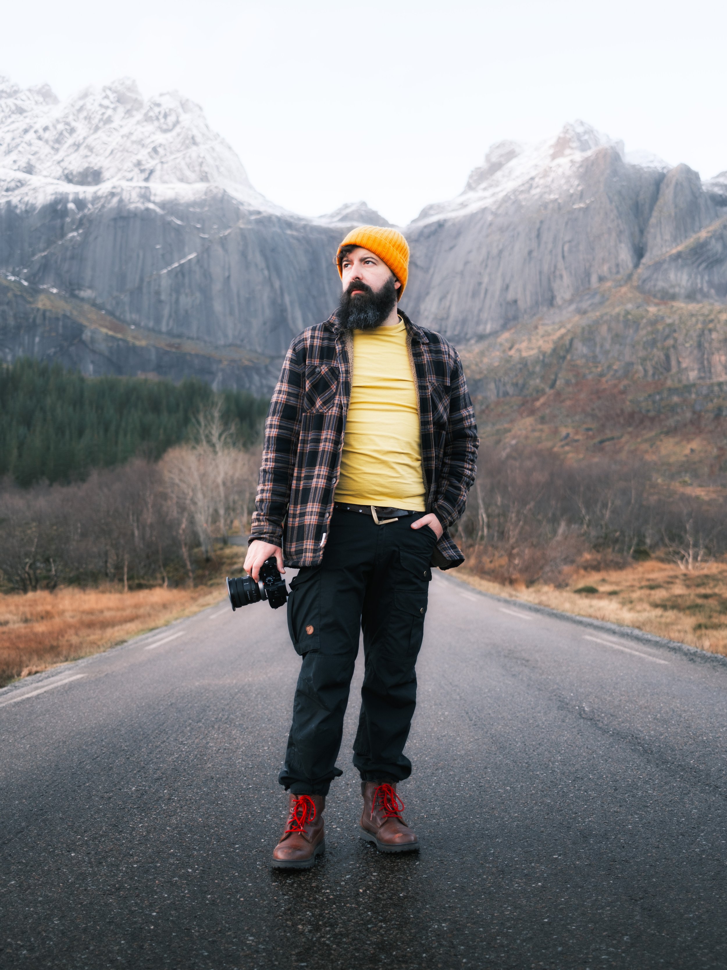 A man with a beard and orange beanie hat standing on an empty road in a mountainous area, holding a camera, with snow-capped mountains in the background.