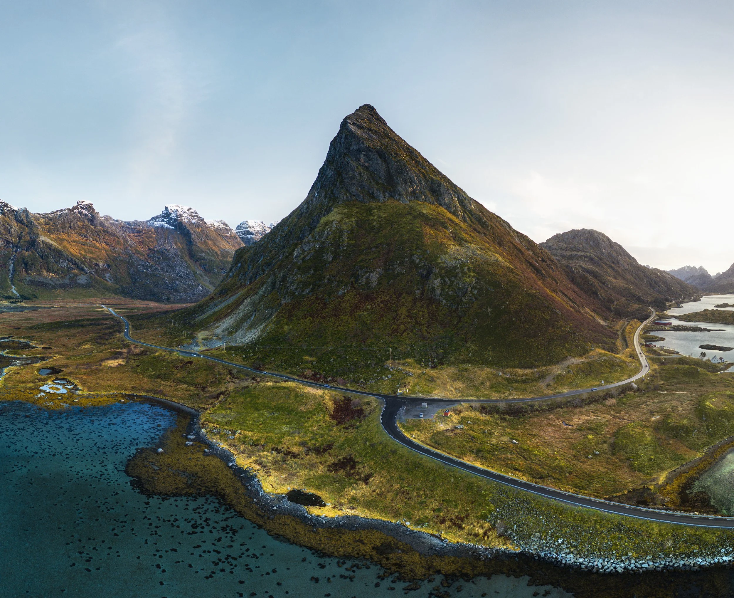 A valley with a prominent mountain peak in the center, surrounded by smaller mountains with snow on their tops, a river winding through the landscape, and a road with a few vehicles.
