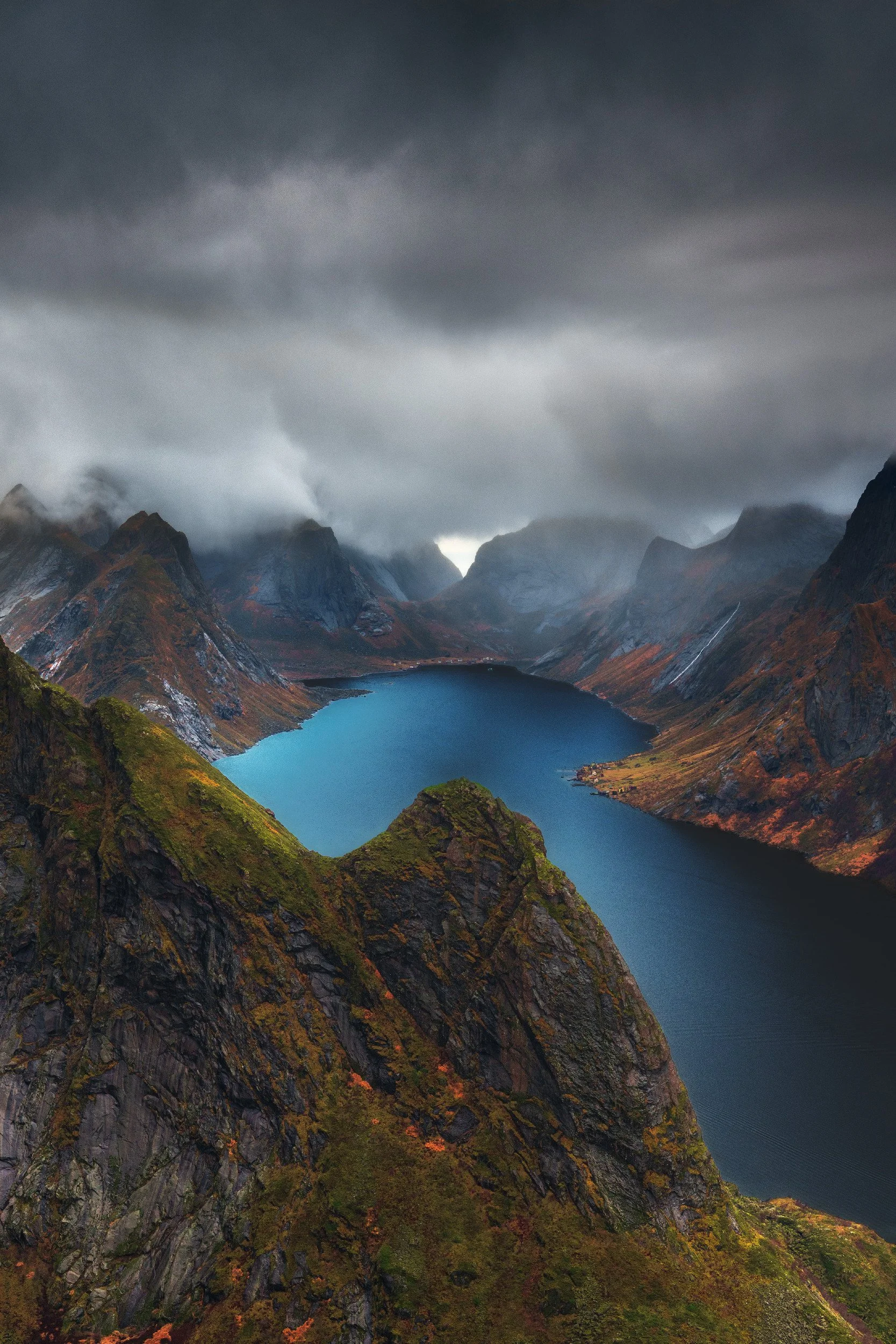 A mountain landscape with dark cloudy skies, rugged mountains, a blue lake, and autumn foliage on the slopes.