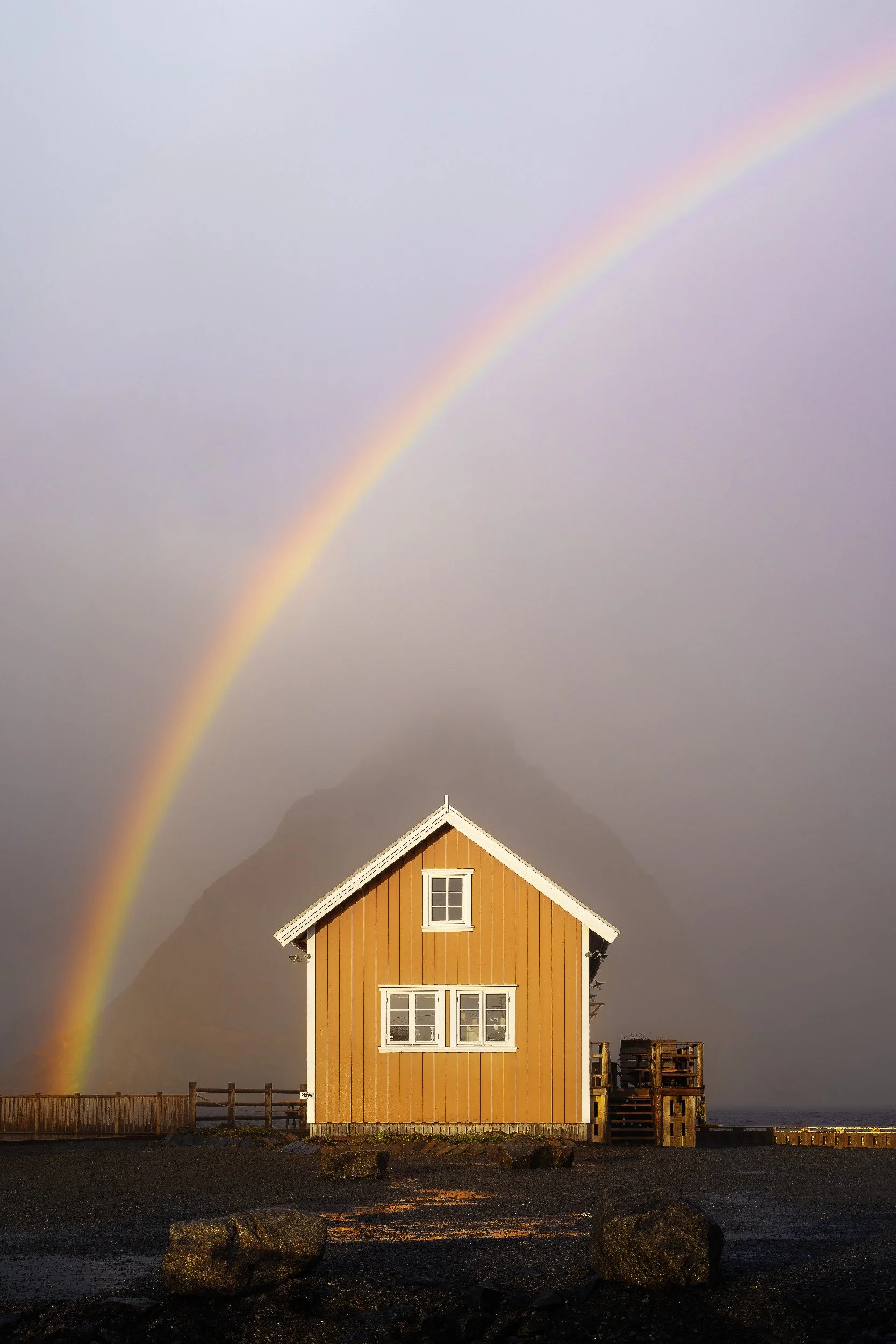 A yellow wooden house with white trim and multiple windows, situated on a concrete surface near a body of water, with rocks in the foreground, a fence, and a rainbow in the cloudy sky.