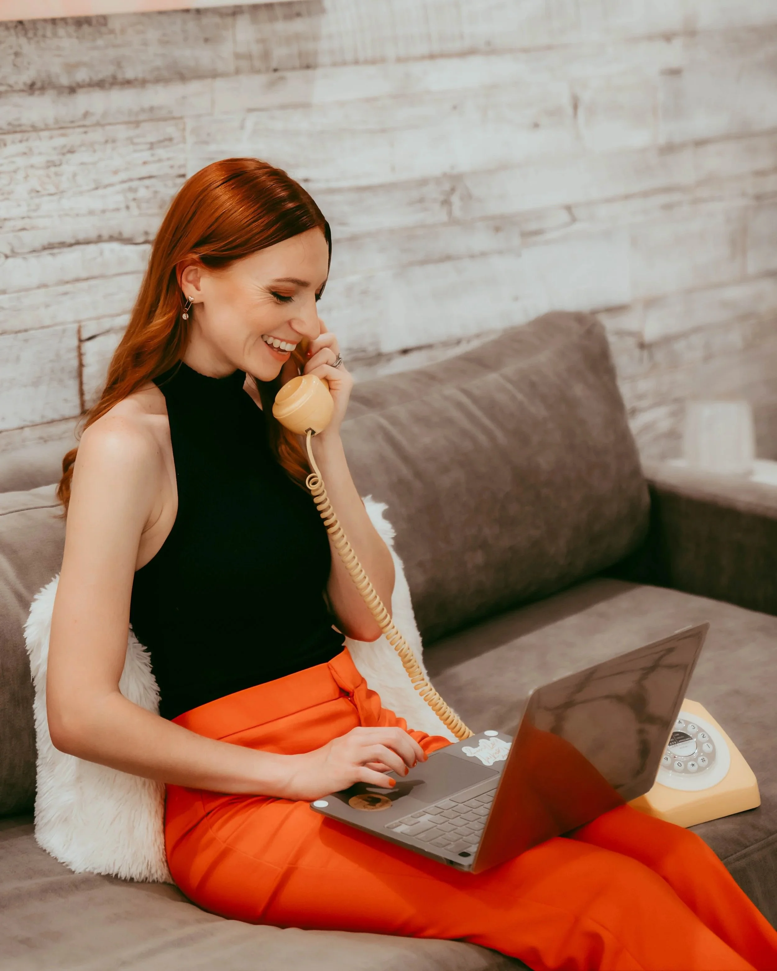 A woman with red hair sitting on a couch, smiling and talking on a vintage rotary phone, using a laptop on her lap.