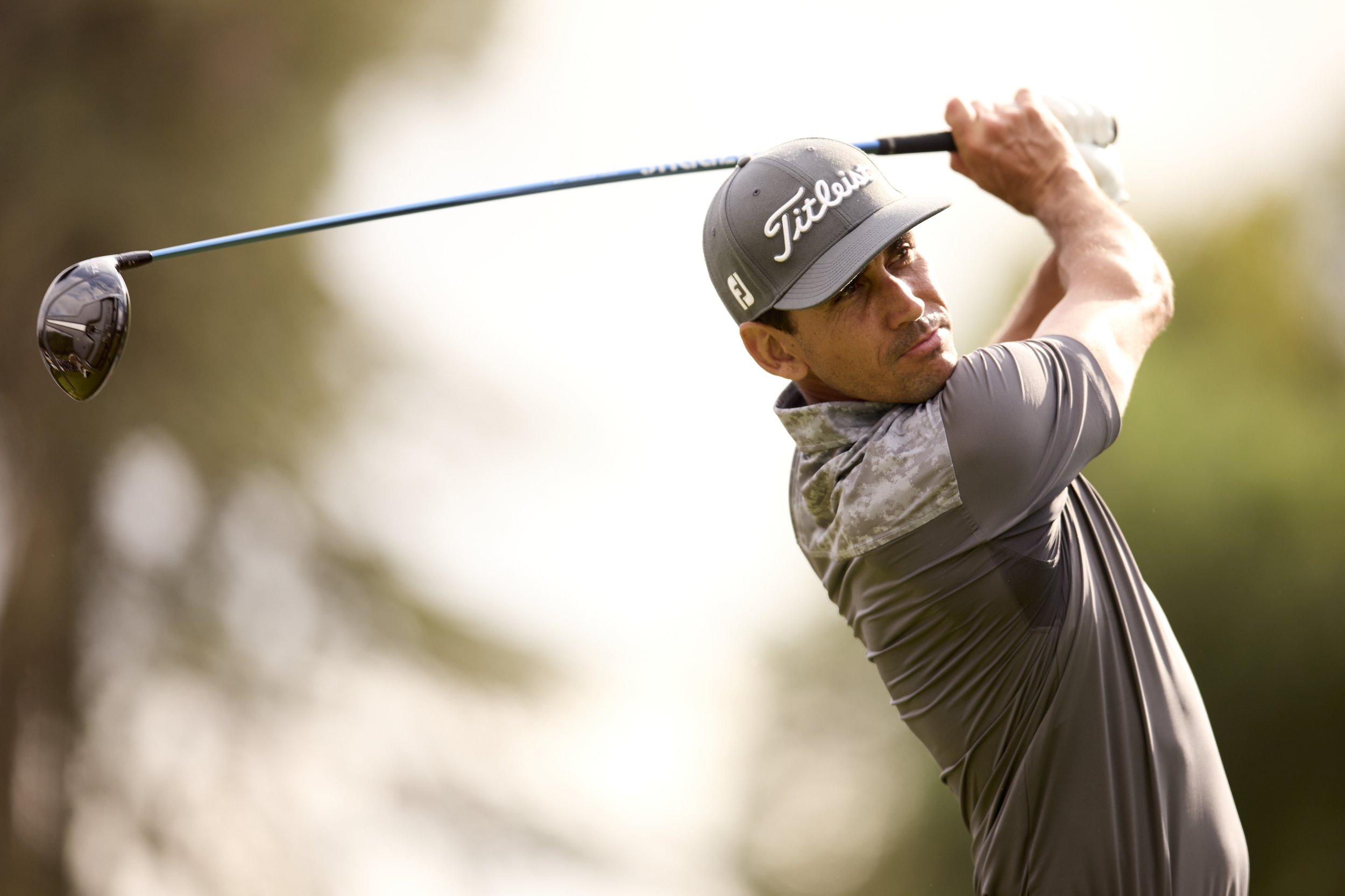 Hombre jugando golf, sosteniendo un palo de golf en movimiento, con fondo de árboles y cielo.