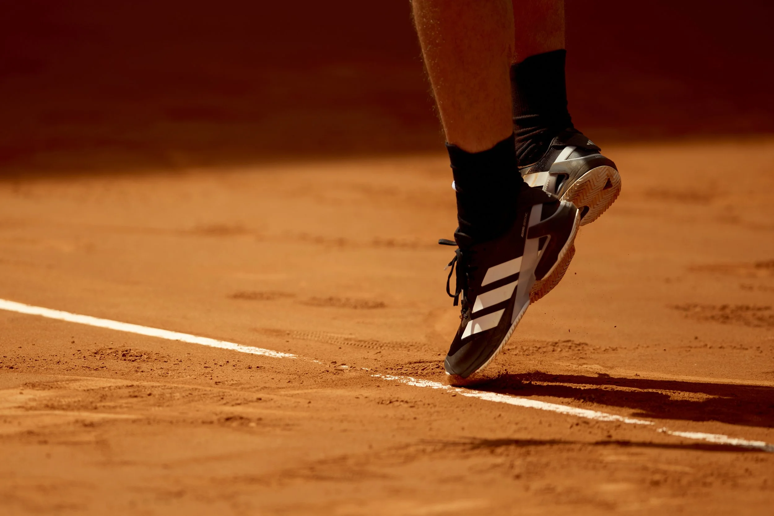 Primer plano de unos zapatos de tenis en movimiento sobre una cancha de arcilla, con líneas blancas. La cancha es de color marrón y la persona lleva pantalones oscuros.