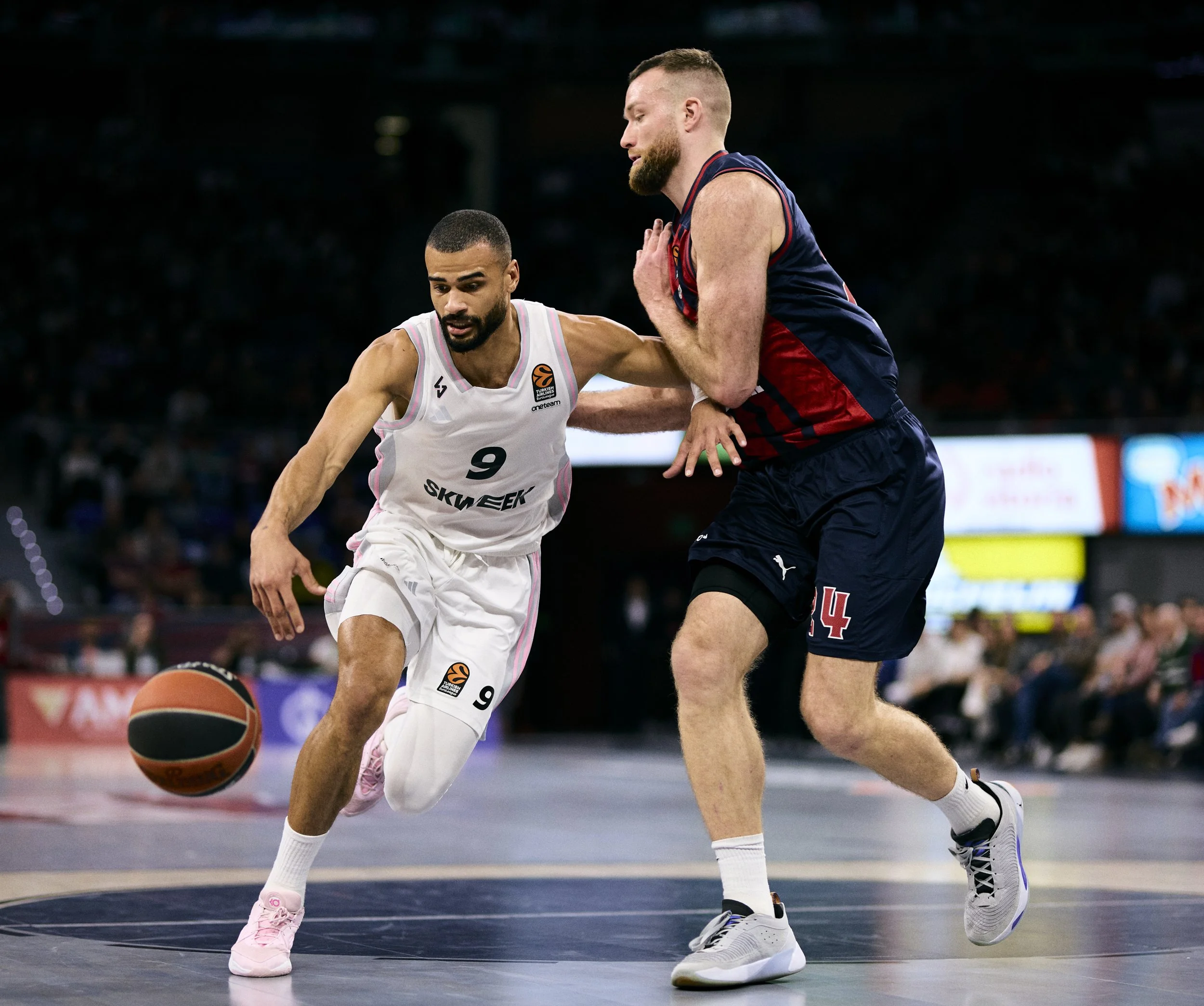 Dos jugadores de baloncesto en acción durante un partido. Uno de ellos, con camiseta blanca y número 9, está controlando la pelota, mientras que el otro, con camiseta azul y número 14, intenta defenderlo.
