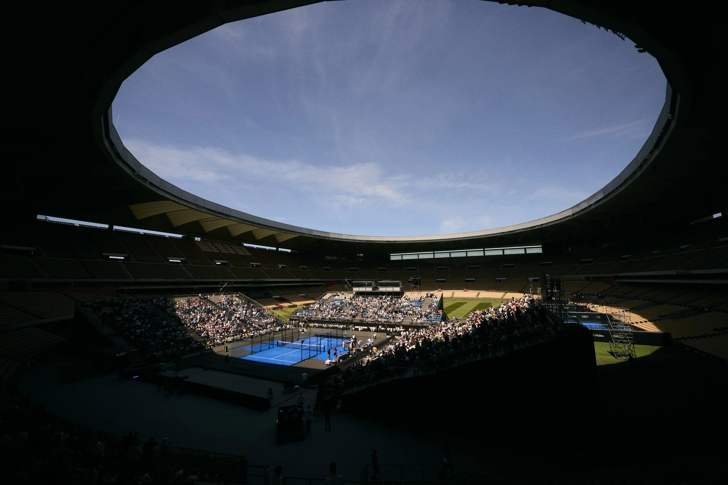 Vista interior de un estadio con cancha de tenis y espectadores, con cielo despejado visible a través de un óvalo en el techo.