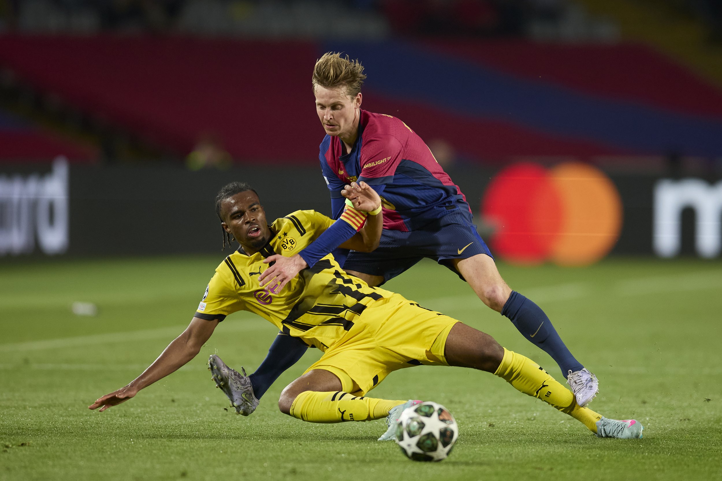 Dos futbolistas en acción durante un partido, uno usando uniforme amarillo y el otro uniforme azul y rojo, luchando por la pelota en el campo.