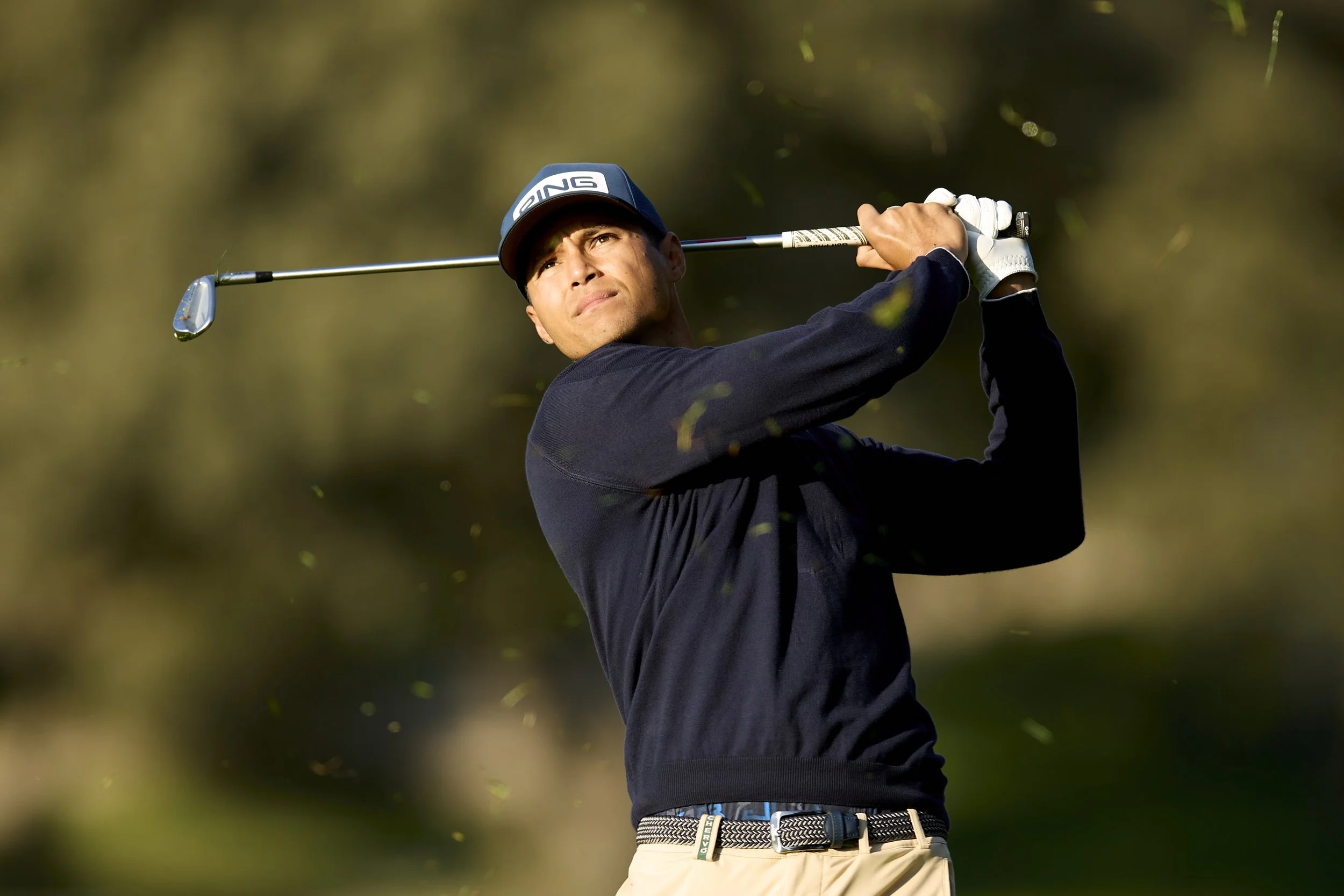 Un hombre jugando golf, sosteniendo un palo en posición de impacto, con fondo borroso de naturaleza.