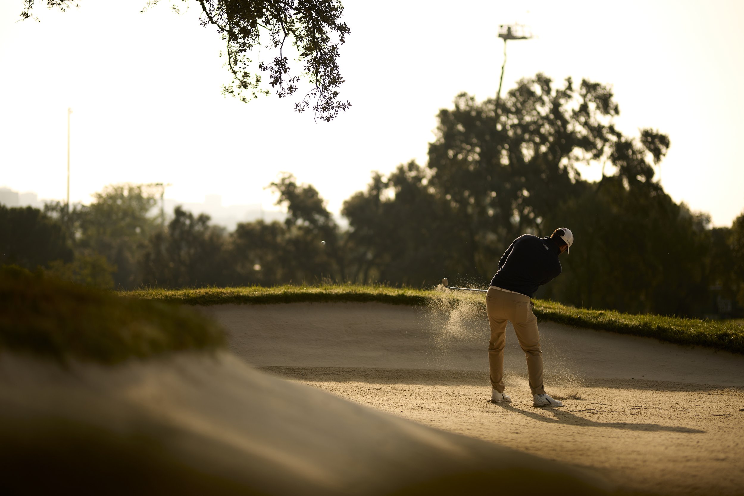 Jugador de golf en un campo de golf, golpeando la pelota desde la arena en un entorno al atardecer, con árboles y cielo brillante al fondo.