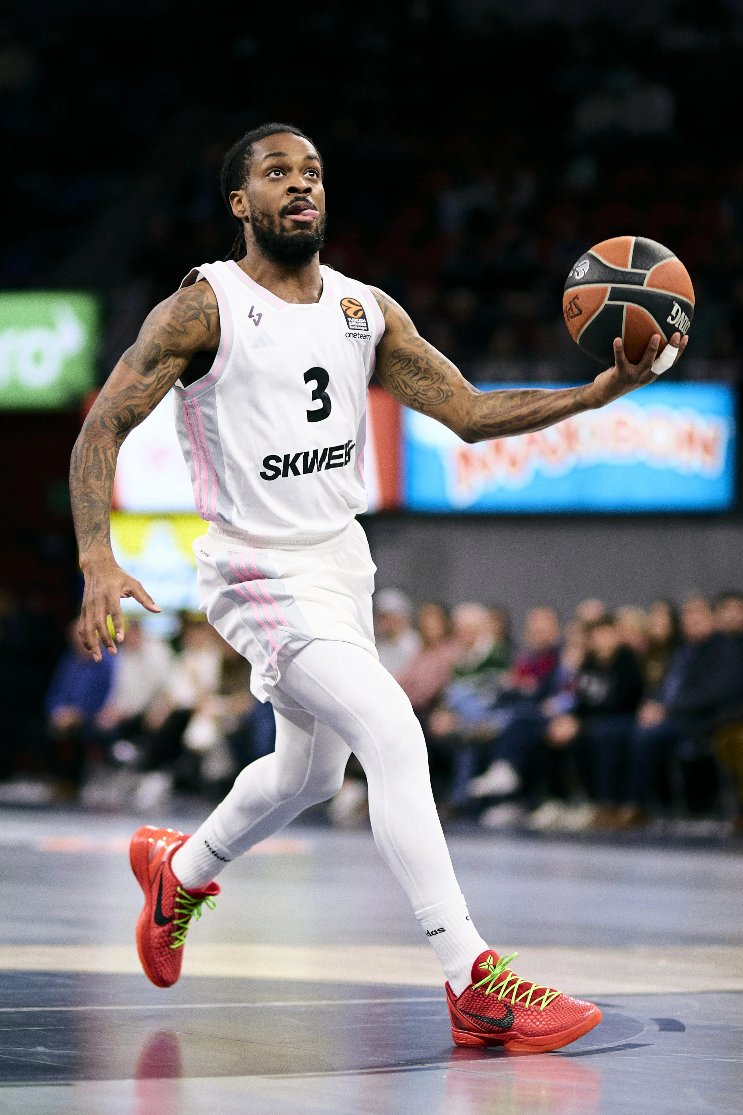 Jugador de baloncesto en acción en la cancha, con uniforme blanco y zapatos rojos, sosteniendo un balón de baloncesto.