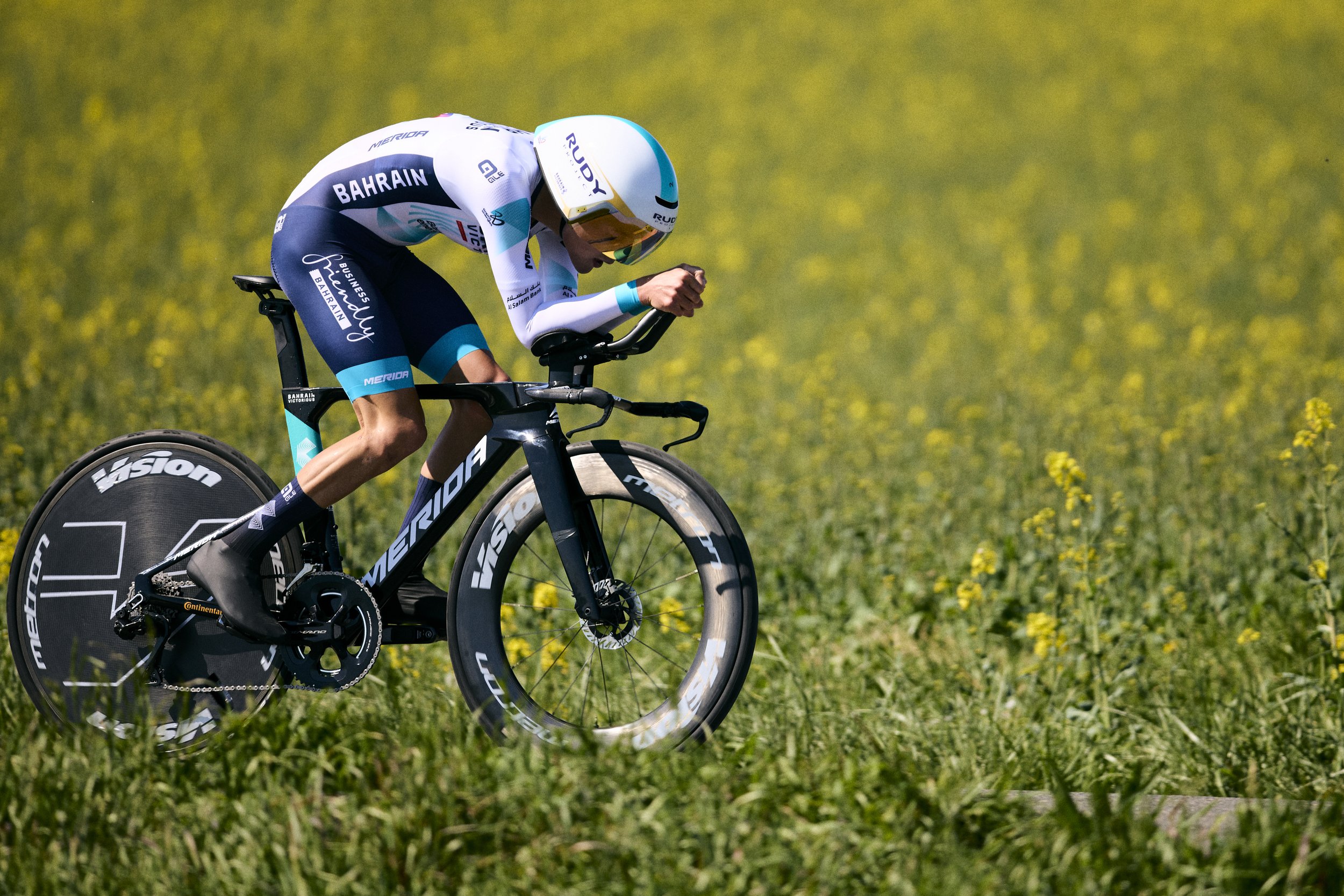 Un ciclista en traje de competición en medio de un campo con flores amarillas en un día soleado, montando una bicicleta de carreras con neumáticos finos.