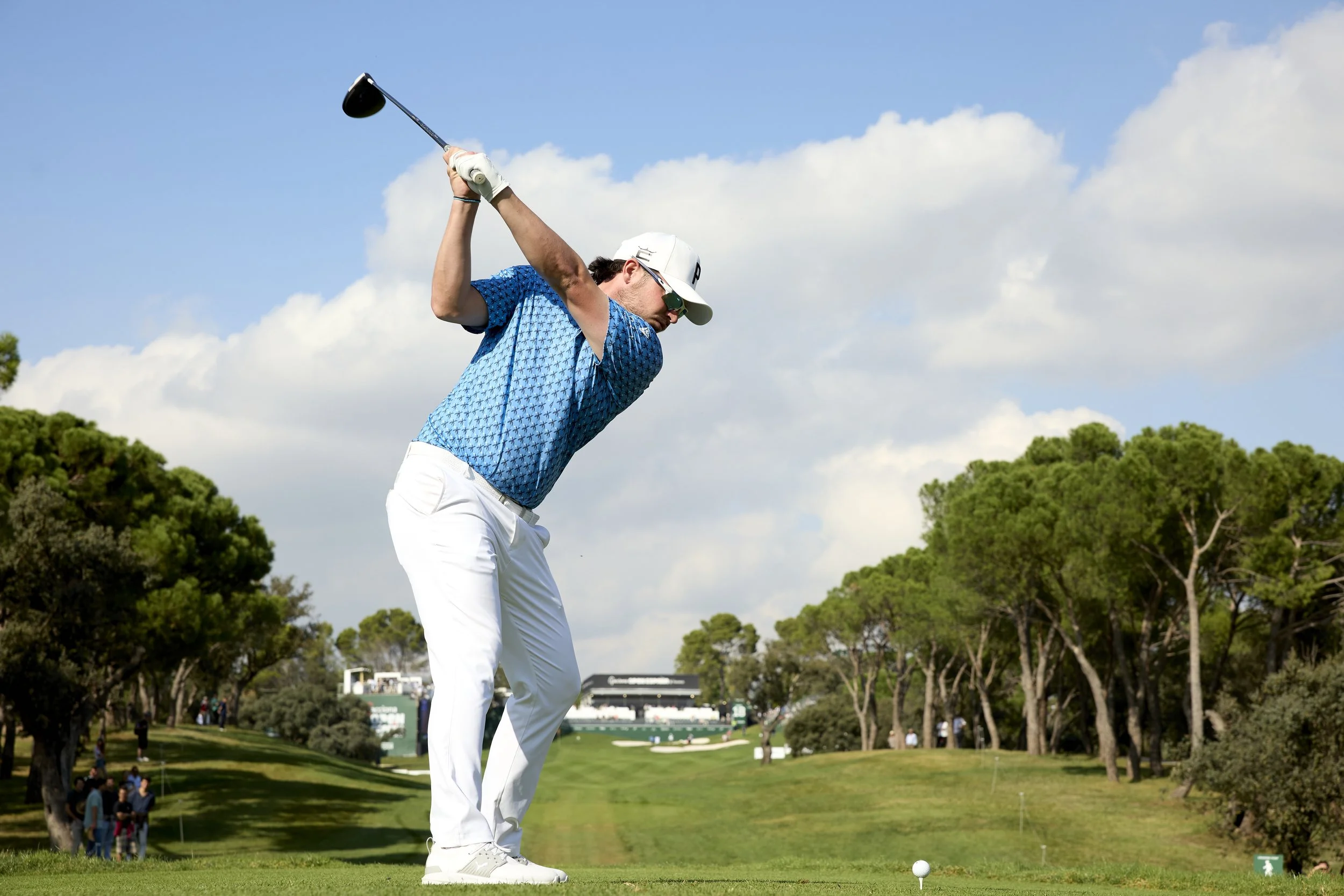 Un golfista masculino en posición de golpe en un campo de golf, con árboles y espectadores al fondo.