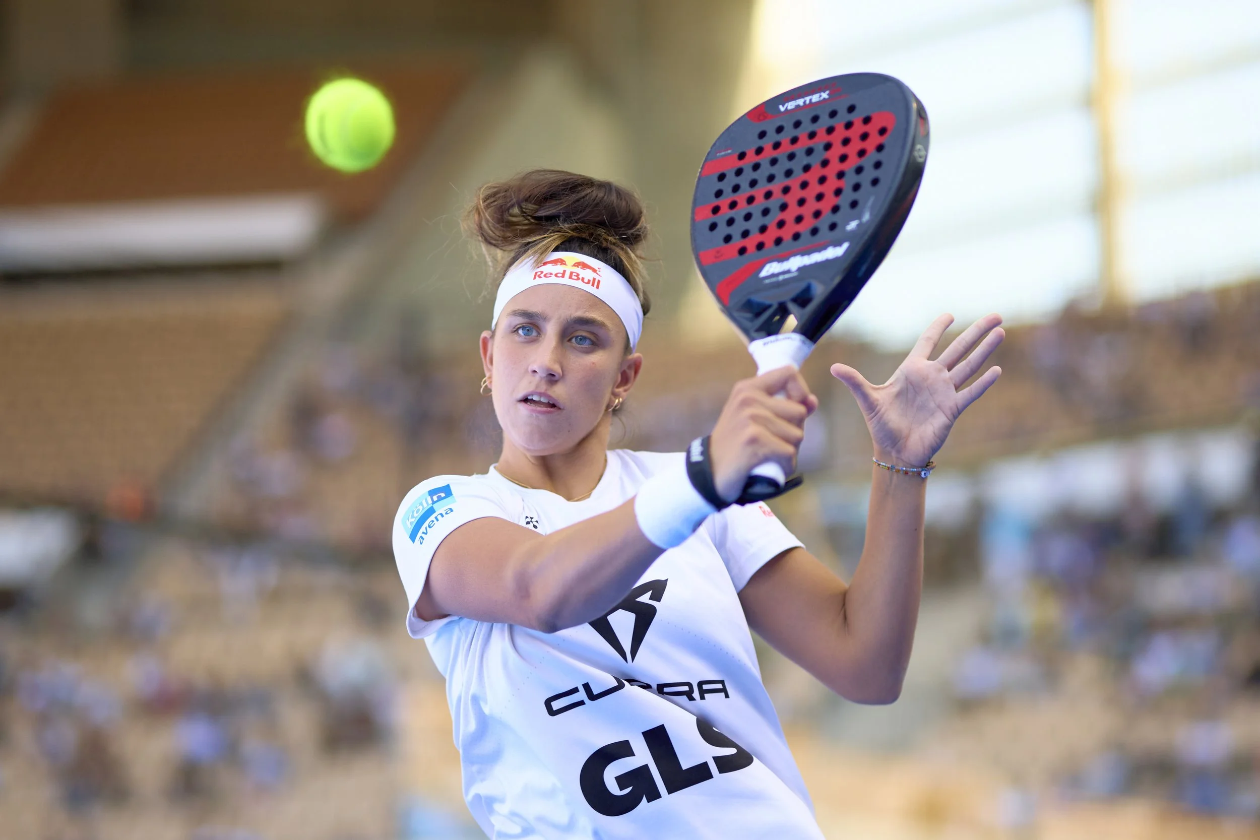 Mujer jugando al pádel en una cancha cerrada, golpeando la pelota con una pala, vestida con ropa deportiva blanca y una diadema blanca con el logo de Red Bull.