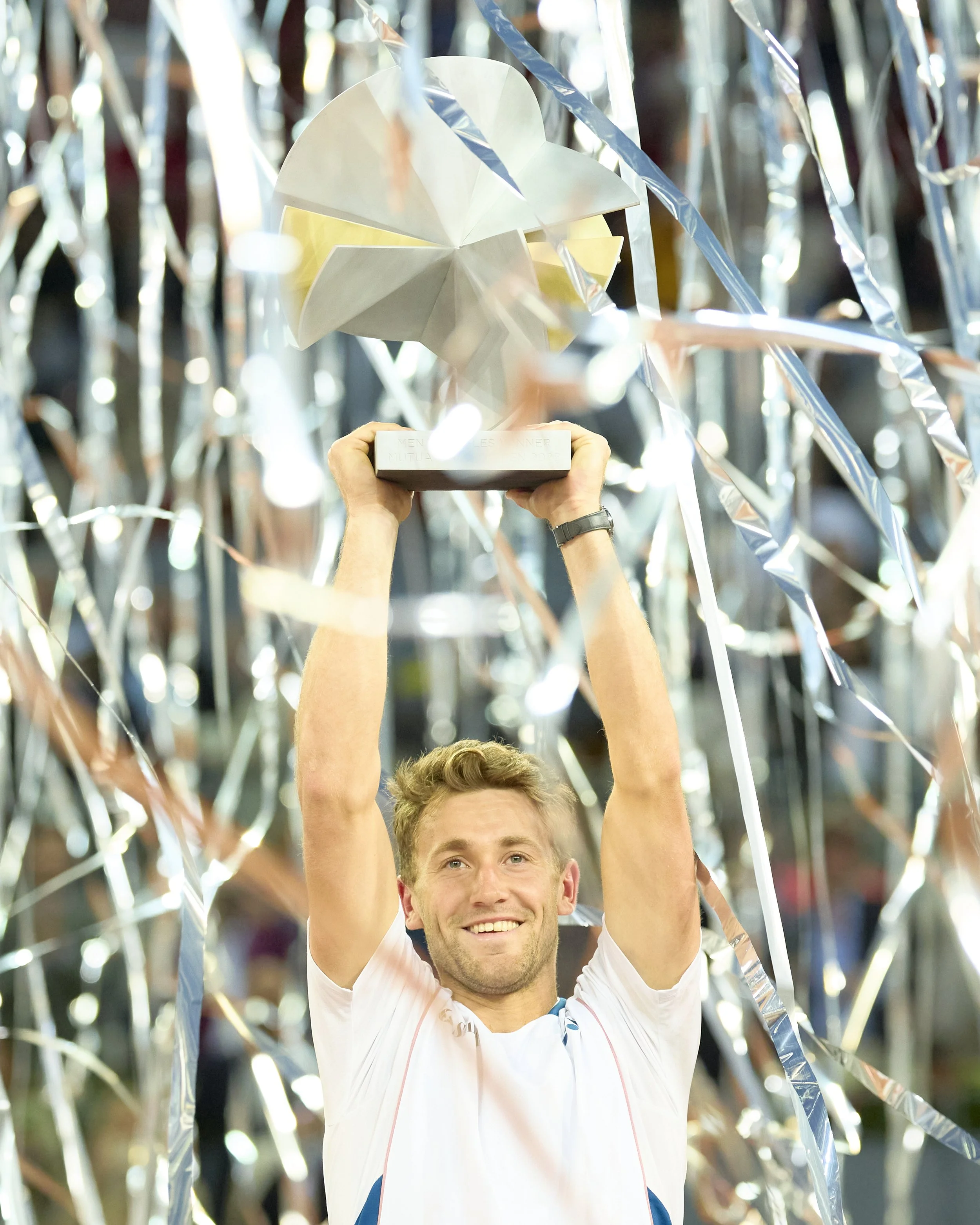 Hombre sonriente levantando un trofeo en un escenario decorado con serpentinas plateadas y papel artesanal.