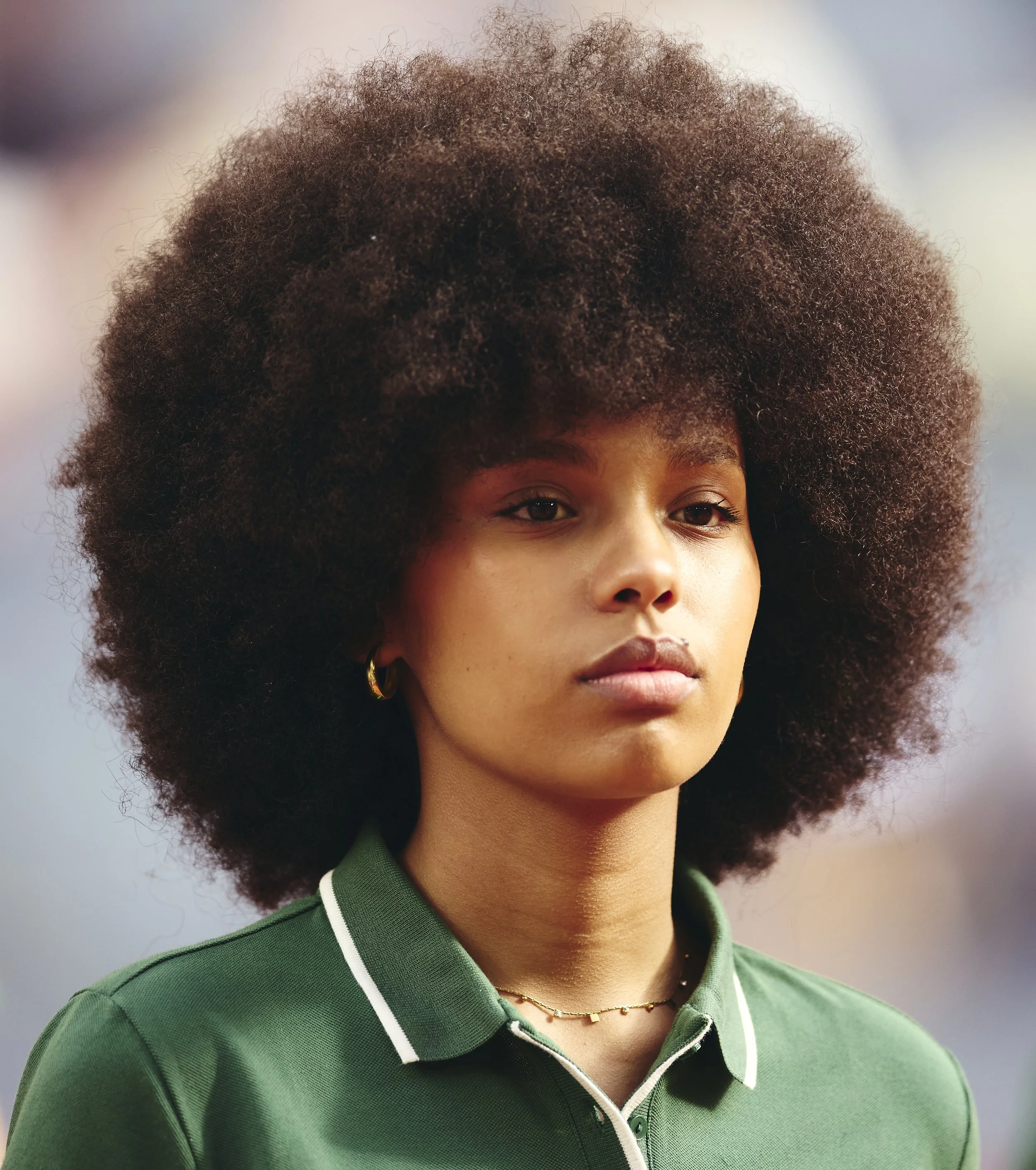 Retrato de una joven con cabello rizado y afro, usando una camiseta deportiva verde con cuello blanco y pequeños detalles dorados, en un fondo desenfocado.