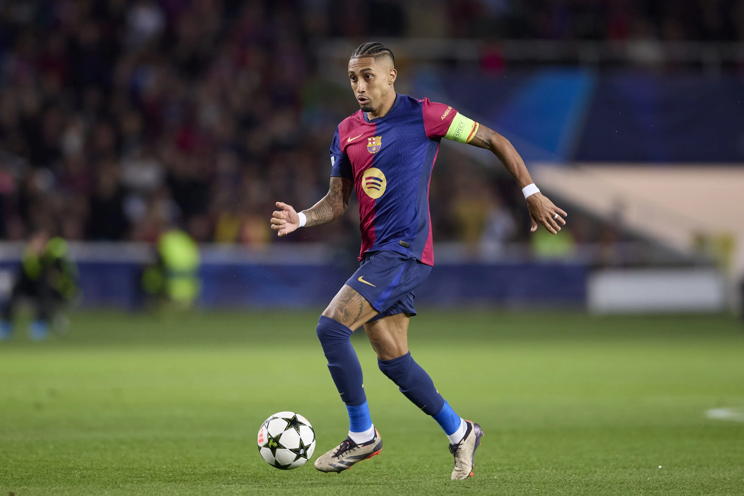 Jugador de fútbol con uniforme del FC Barcelona en el campo, controlando el balón durante un partido, con público en las gradas al fondo.