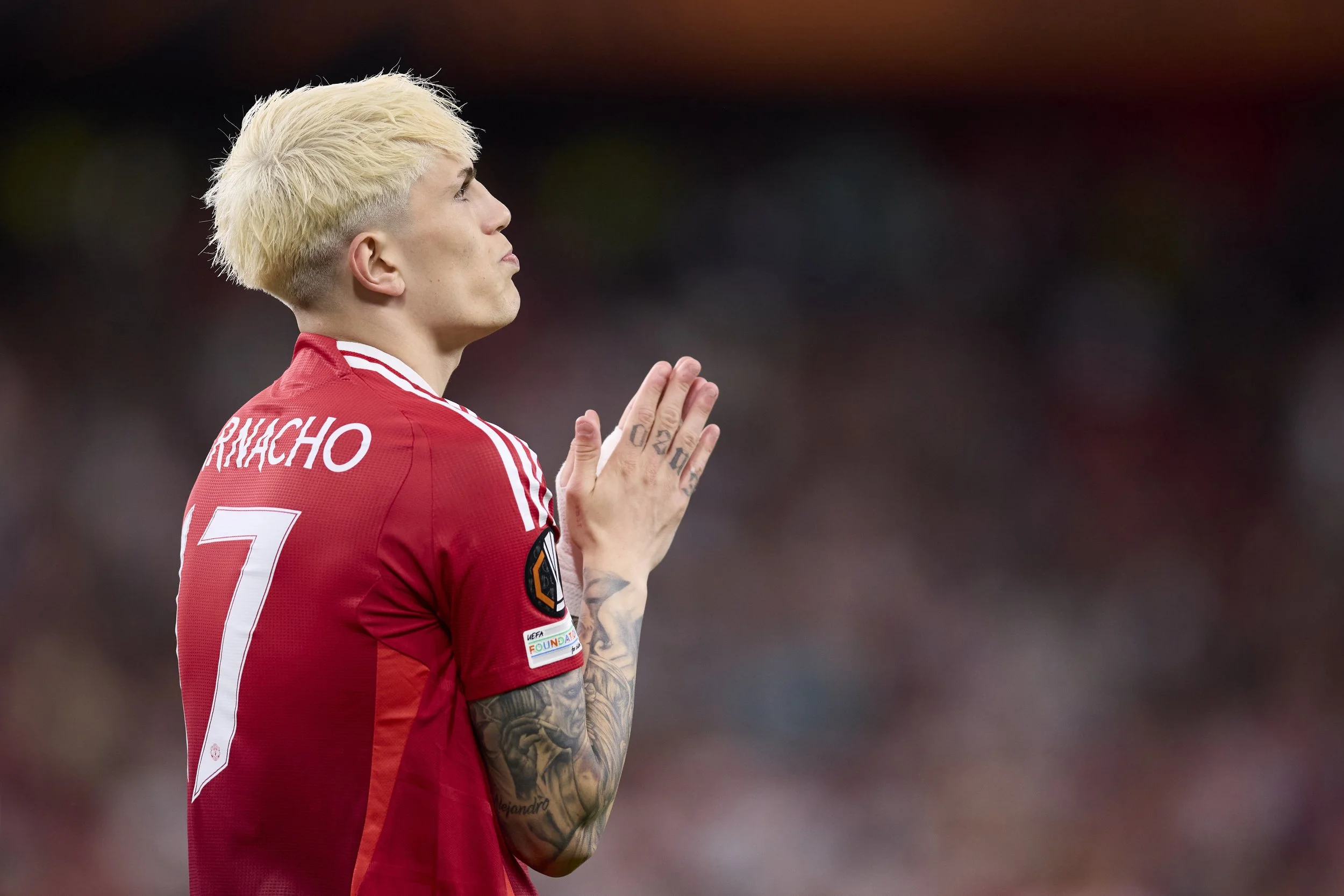 Jugador de fútbol con camiseta roja con el número 7 y el nombre 'RICHACHO' en la espalda, en una posición de oración o agradecimiento en el campo de fútbol.
