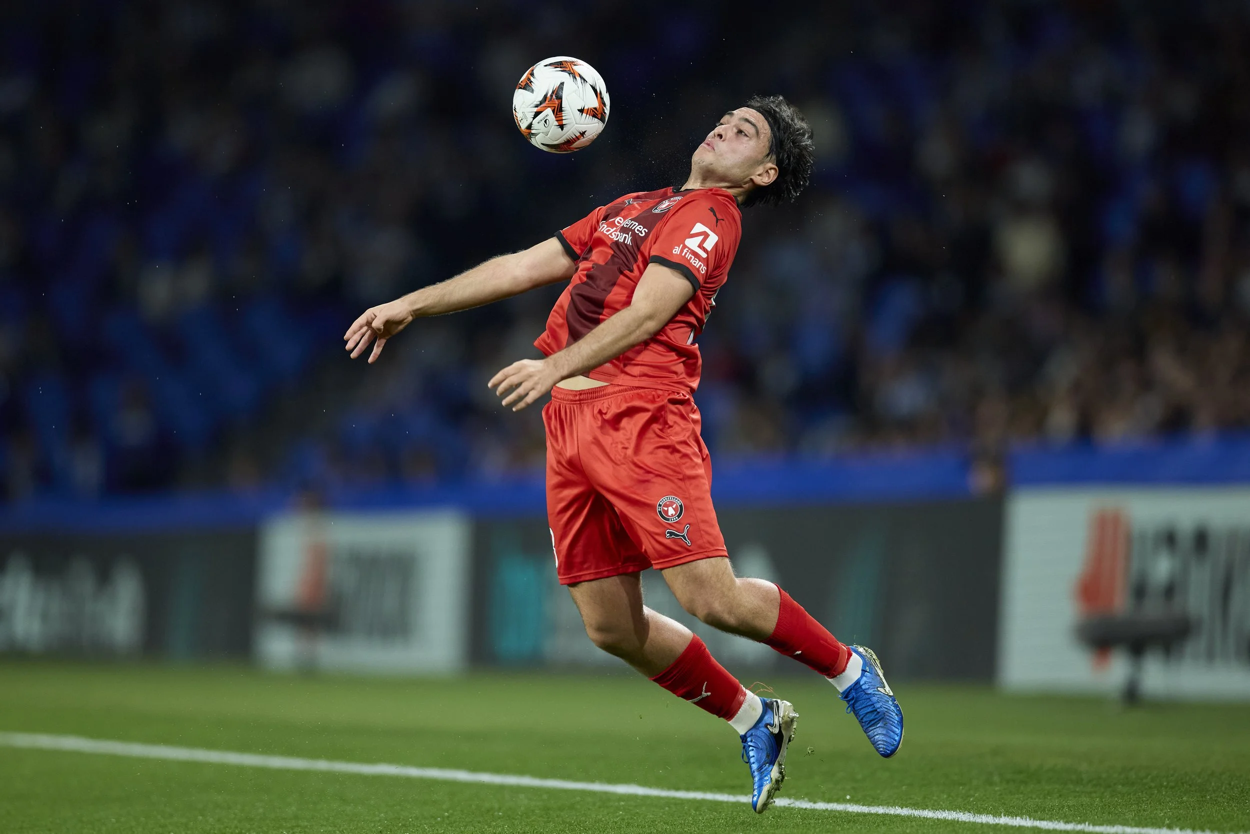 Jugador de fútbol en uniforme rojo, saltando para cabecear un balón en un partido.